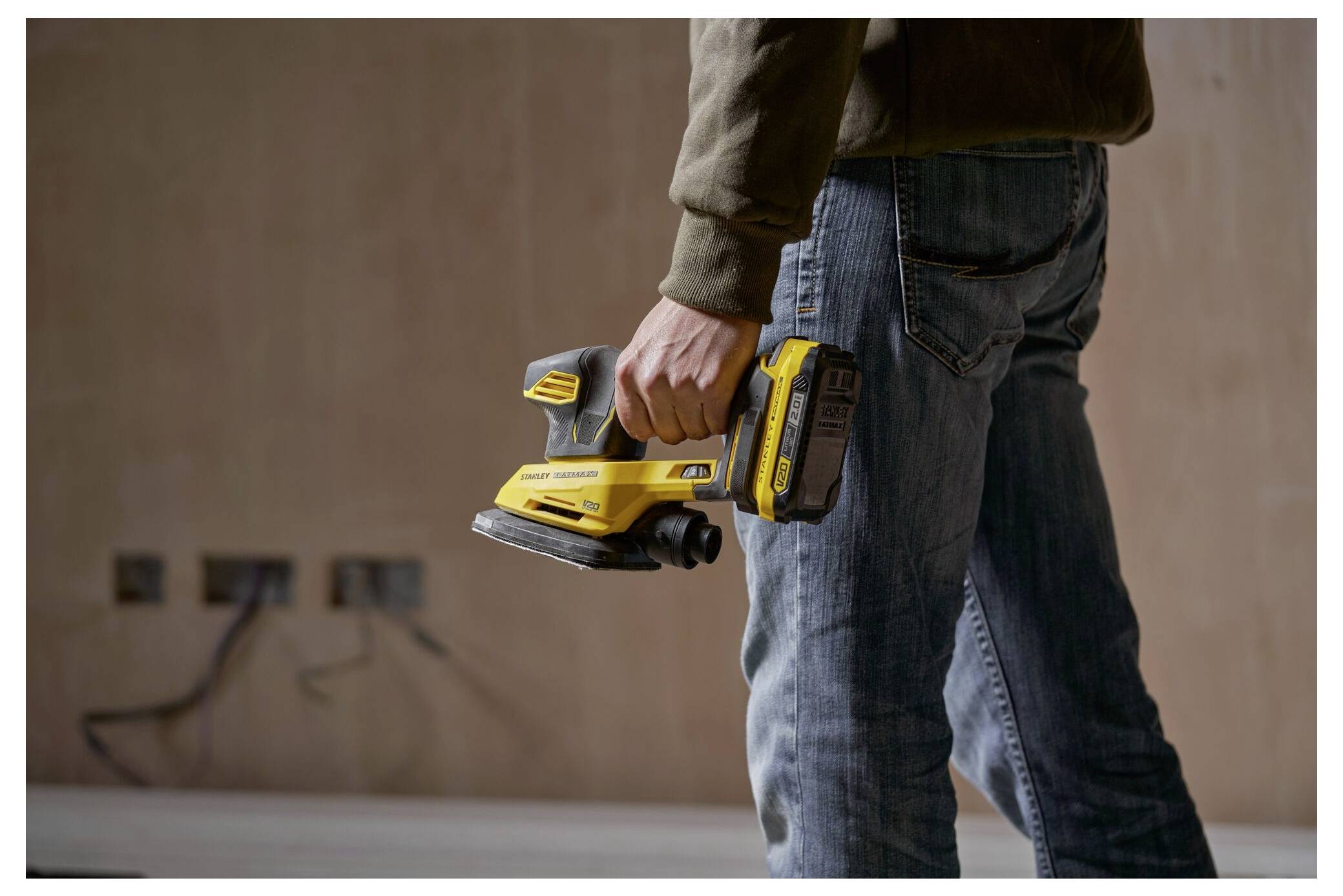 Eine Person hält einen gelben Elektroschleiffer in einem teilweise renovierten Raum mit freigelegten Steckdosen in der Wand.