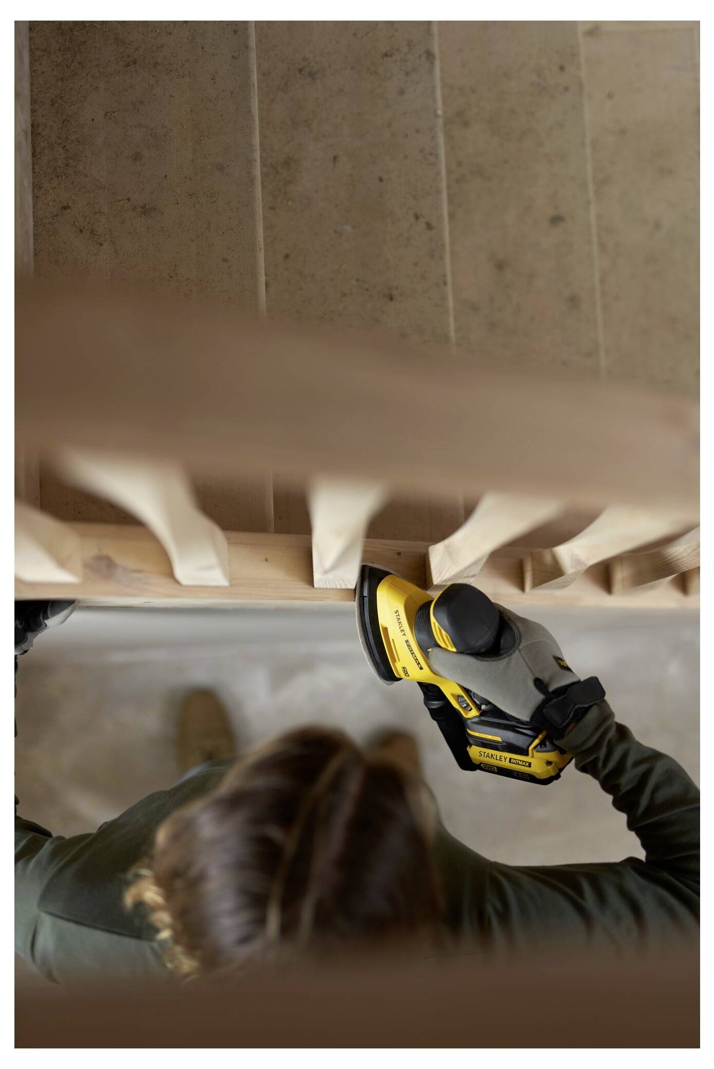 Person mit einem gelben Elektroschleiffer beim Glätten eines hölzernen Treppengeländers. Ansicht von oben.