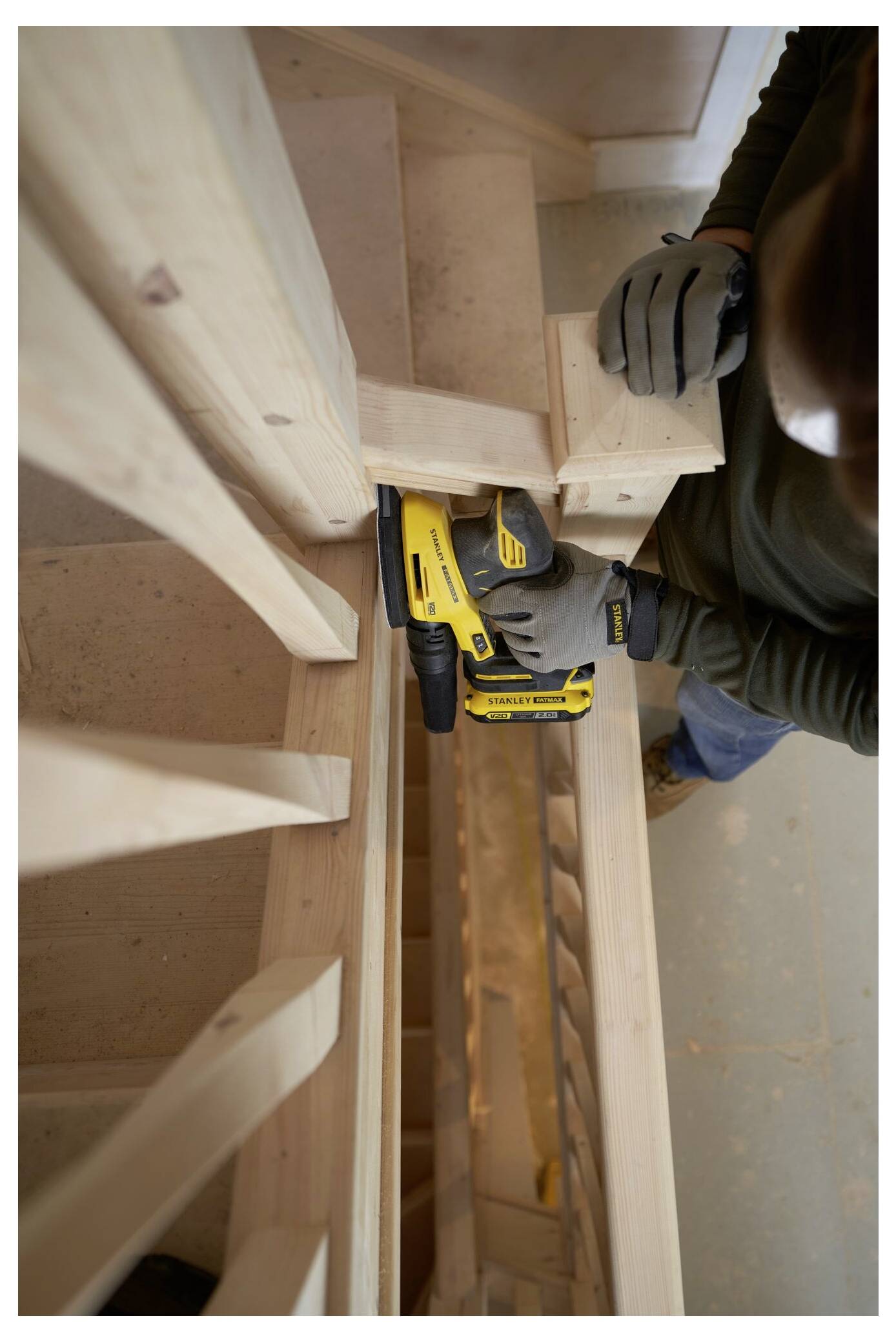 Eine Person mit Handschuhen verwendet ein gelbes Elektrowerkzeug, um hölzerne Treppen zusammenzubauen oder zu reparieren, und konzentriert sich dabei auf die Befestigung des Geländers.