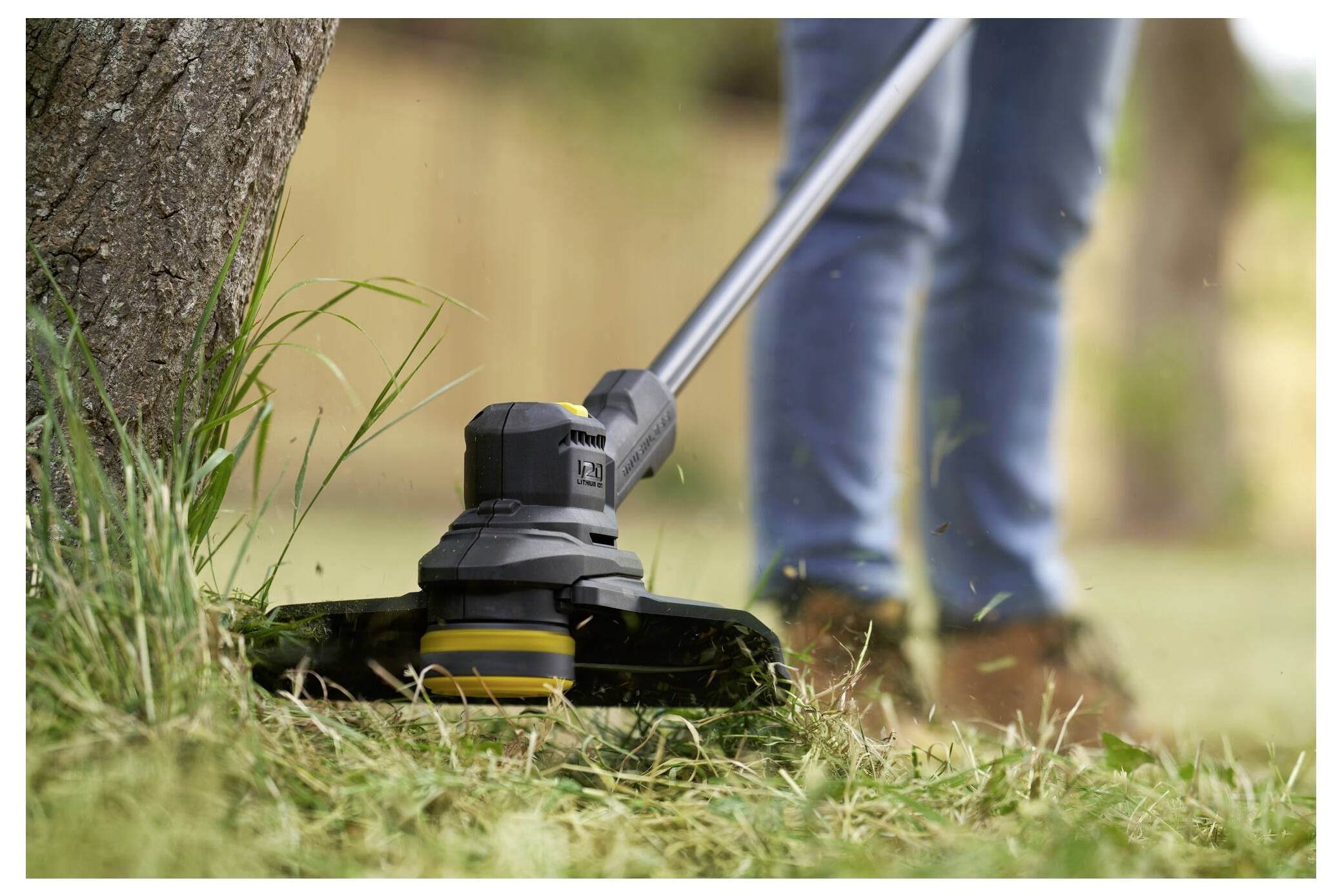 Eine Person, die einen Rasentrimmer in der Nähe eines Baumes benutzt, trägt Jeans und Stiefel und schneidet Gras in einer Außenumgebung.