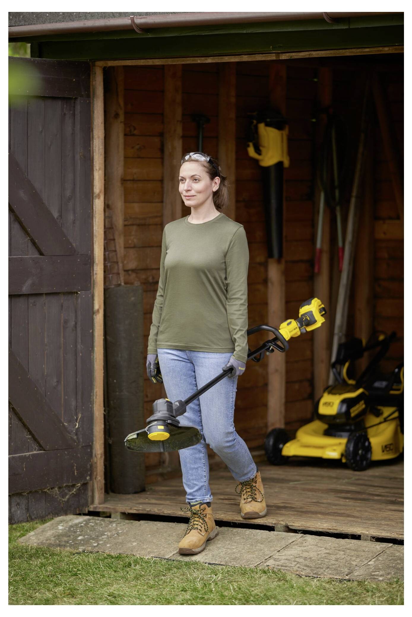 Eine Person in legerer Outdoor-Kleidung und Handschuhen verlässt einen Holzschuppen und hält einen gelb-schwarzen Rasentrimmer in der Hand.
