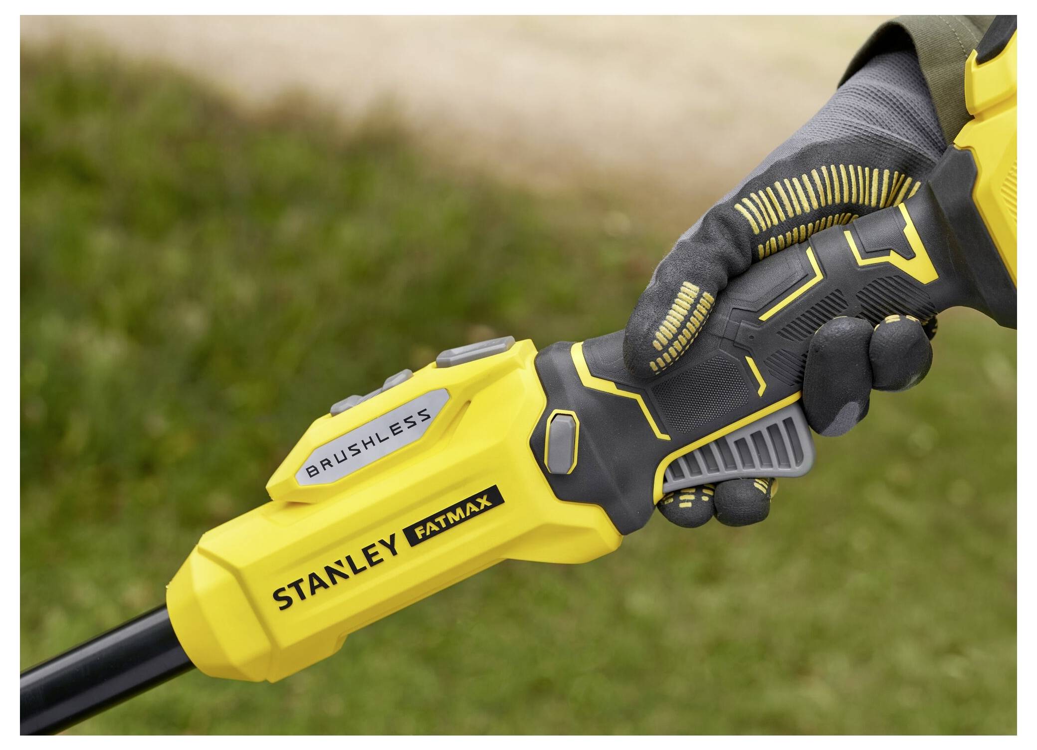 Eine behandschuhte Hand, die ein gelbes und schwarzes Stanley Fatmax bürstenloses Werkzeug greift, mit Gras und einem unscharfen Weg im Hintergrund.
