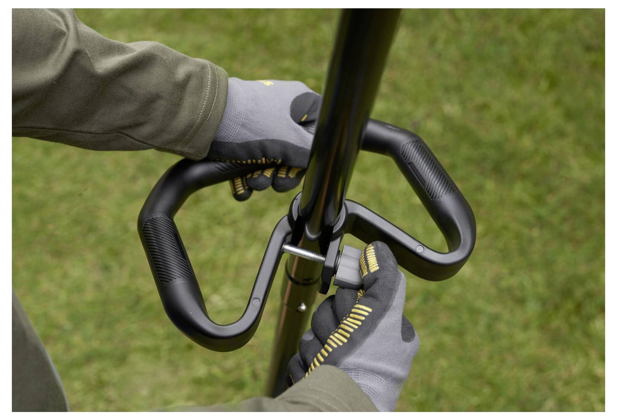 Eine Person, die Handschuhe trägt, stellt den Griff eines Gartengeräts ein und steht auf Gras.
