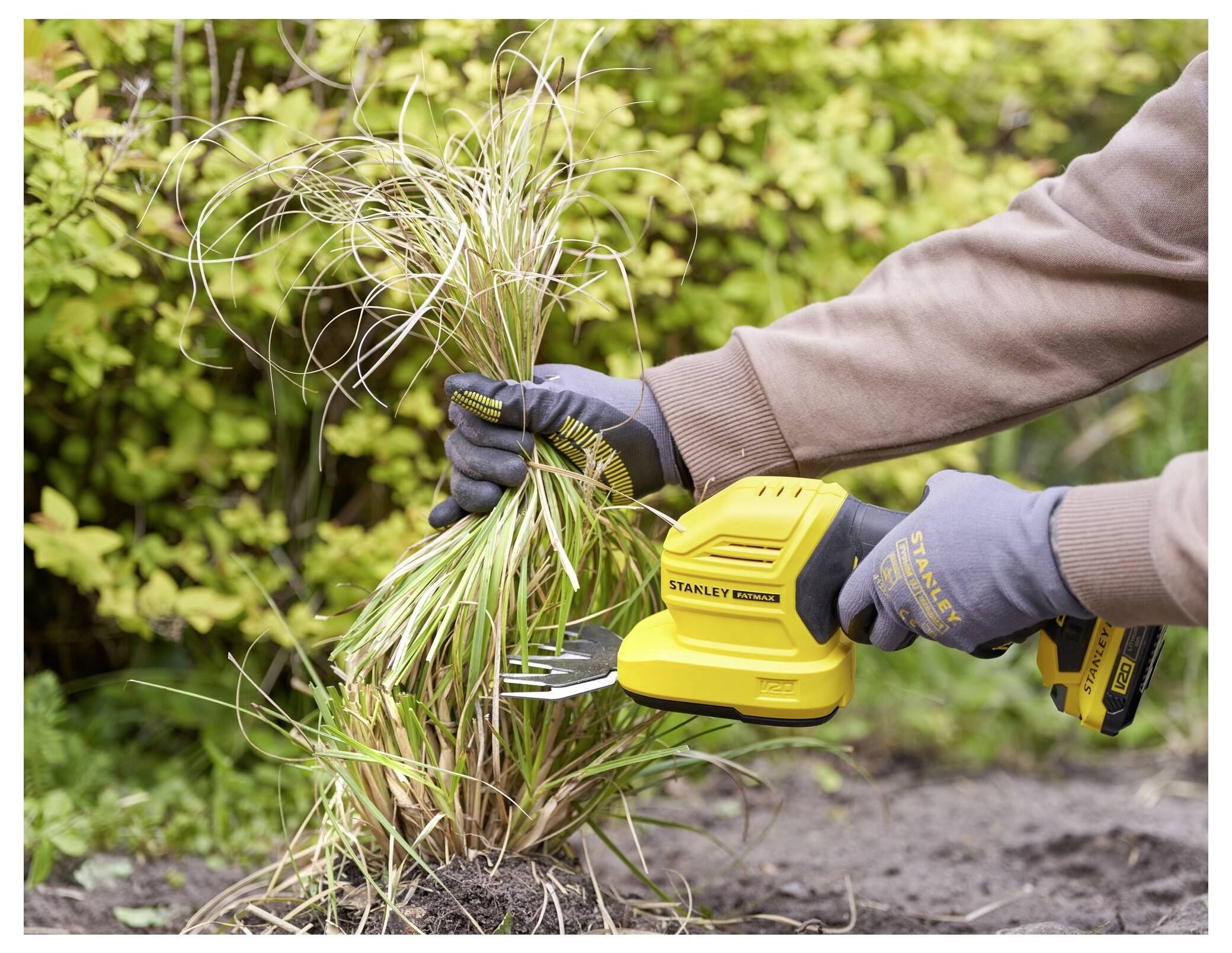 Eine Person, die Handschuhe trägt, schneidet Gras mit einem gelben Elektrowerkzeug in einem Garten, umgeben von grünem Laub.