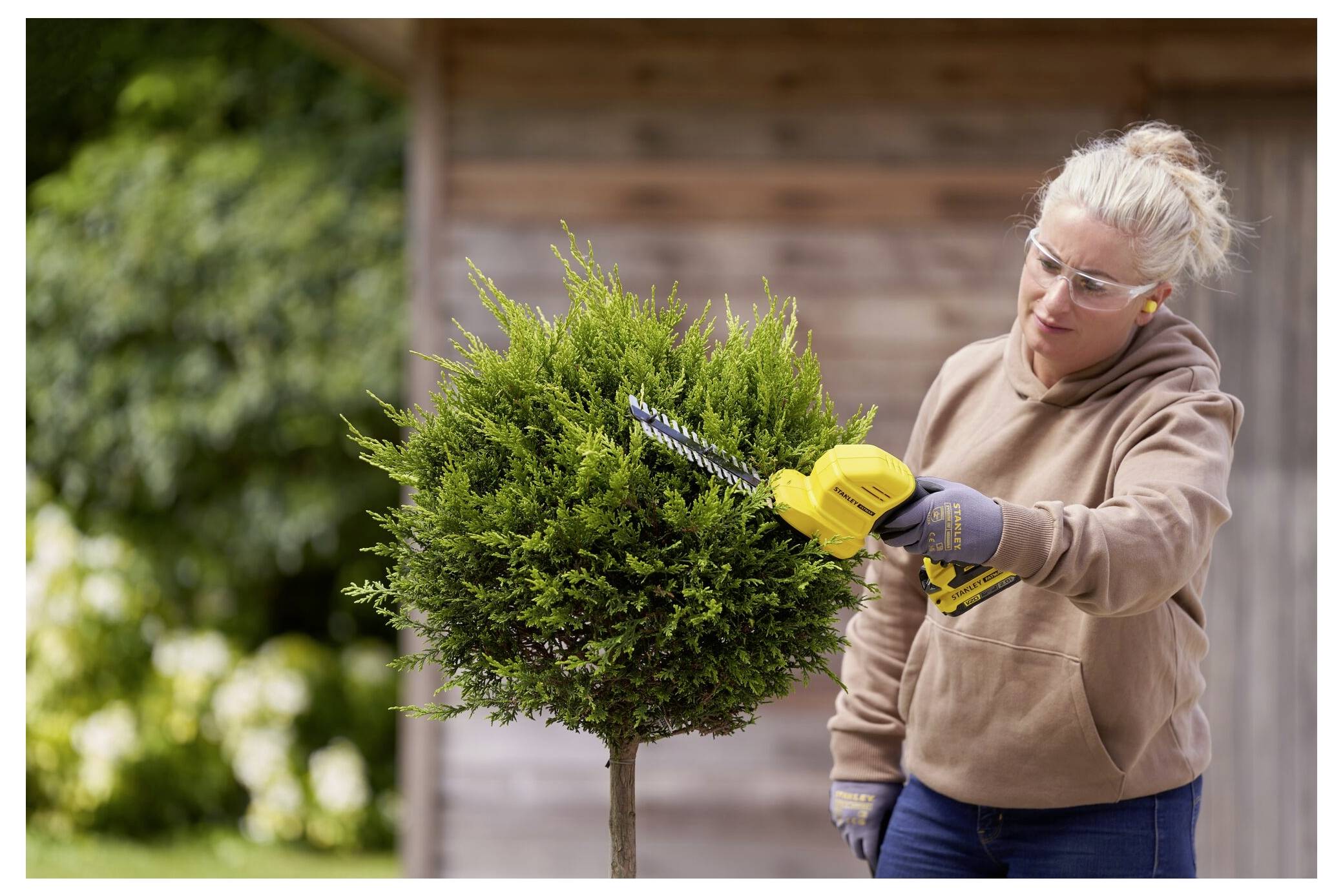 Eine Person schneidet einen kugelförmigen Strauch mit einer elektrischen Heckenschere in einer Gartenanlage, wobei sie eine Schutzbrille und Handschuhe trägt.