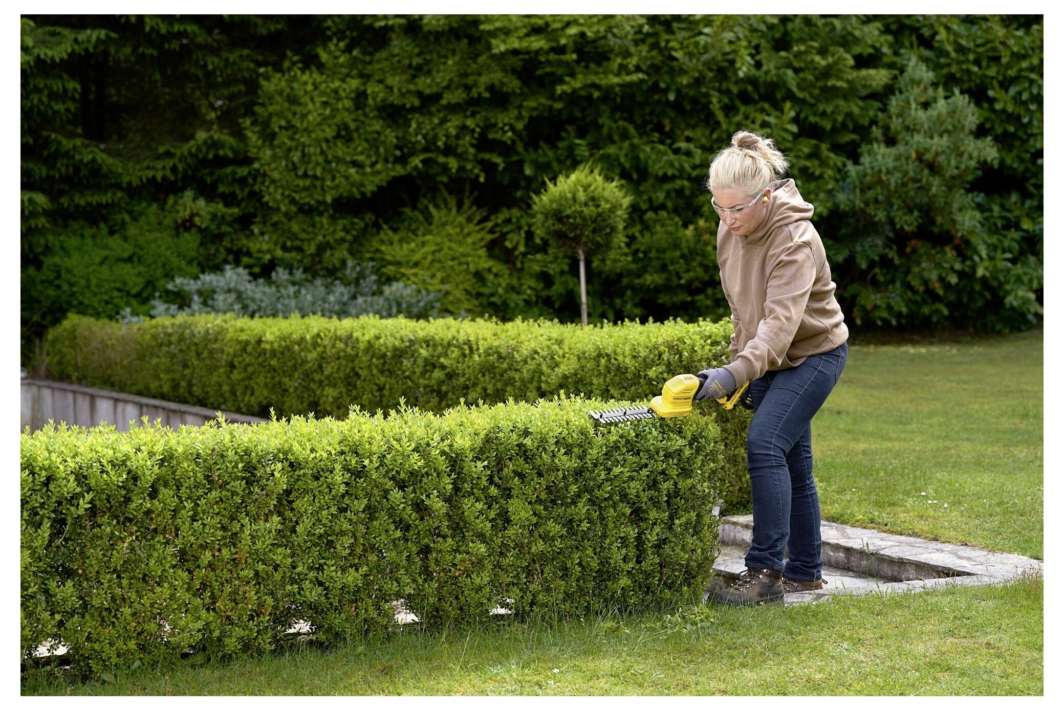 Eine Person, die eine ordentlich geformte Hecke in einem Garten trimmt, trägt Handschuhe und eine Schutzjacke und ist von üppigem Grün umgeben.