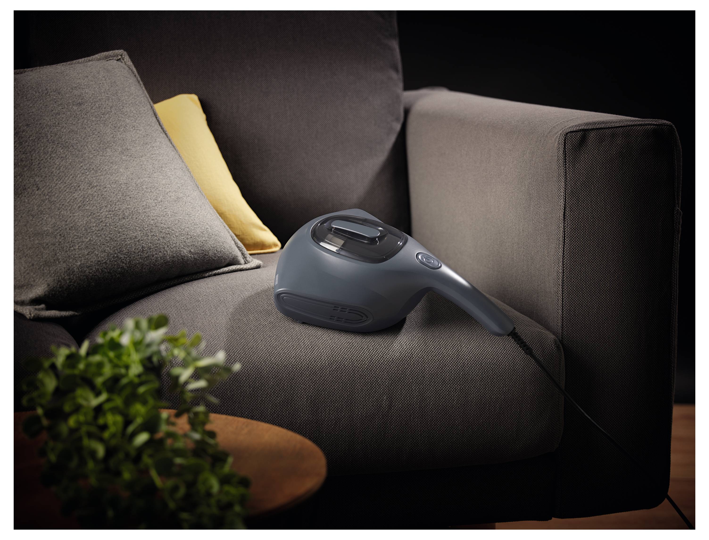 Ein tragbarer Staubsauger ruht auf einem dunkelgrauen Sofa neben gelben und grauen Kissen, mit einer Pflanze, die im Vordergrund sichtbar ist.