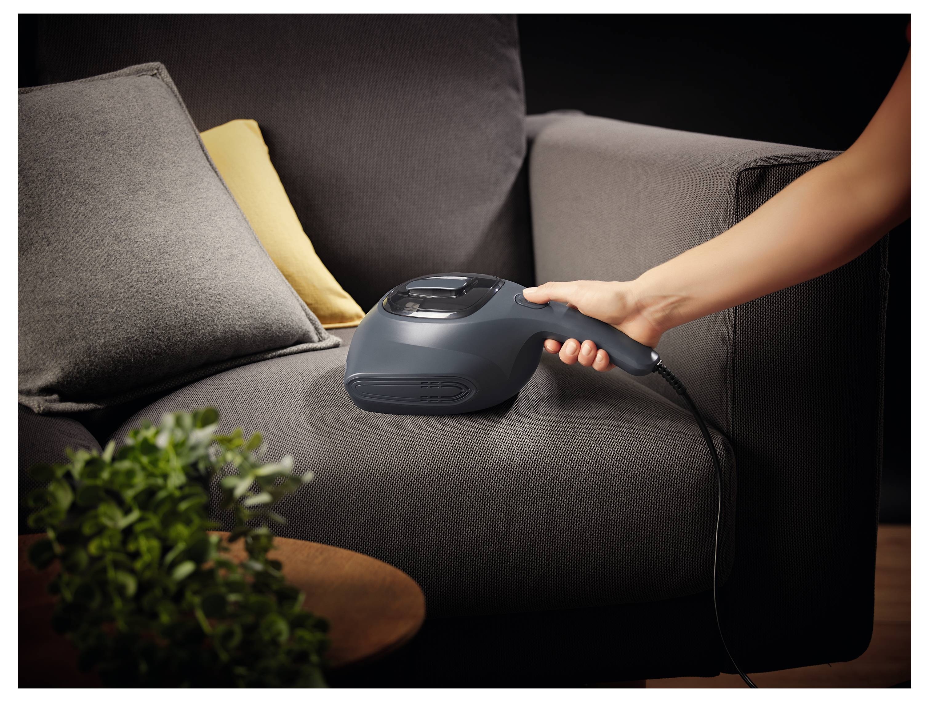 Eine Person benutzt einen tragbaren Staubsauger, um ein dunkelgraues Sofa zu reinigen, auf dem ein beige und gelbes Kissen liegt. Eine kleine Pflanze ist in der Nähe sichtbar.