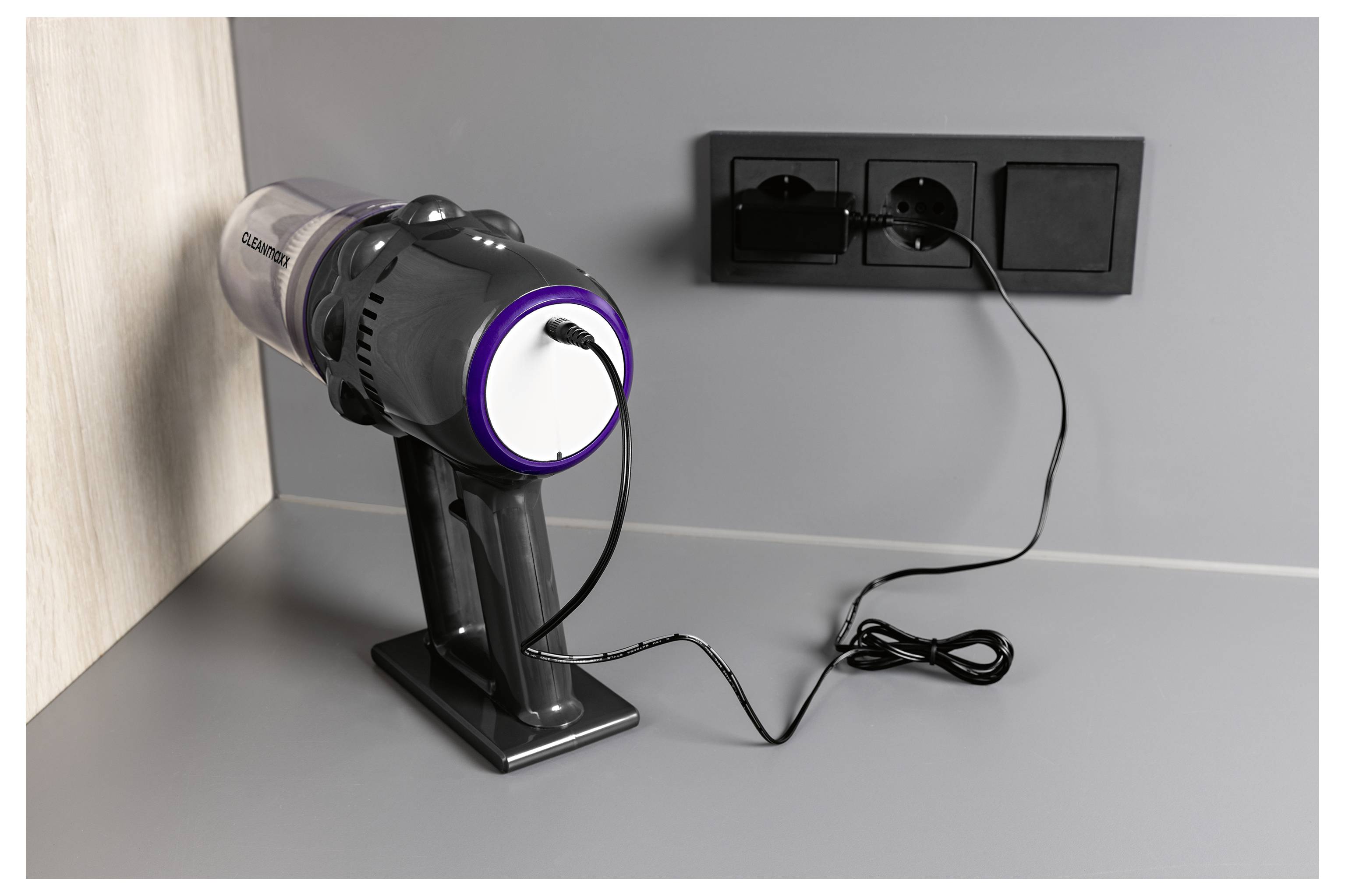 Ein tragbarer Staubsauger ist in eine Wandsteckdose eingesteckt und lädt auf einer Arbeitsplatte mit grauer Oberfläche und hellem Holzhintergrund auf.