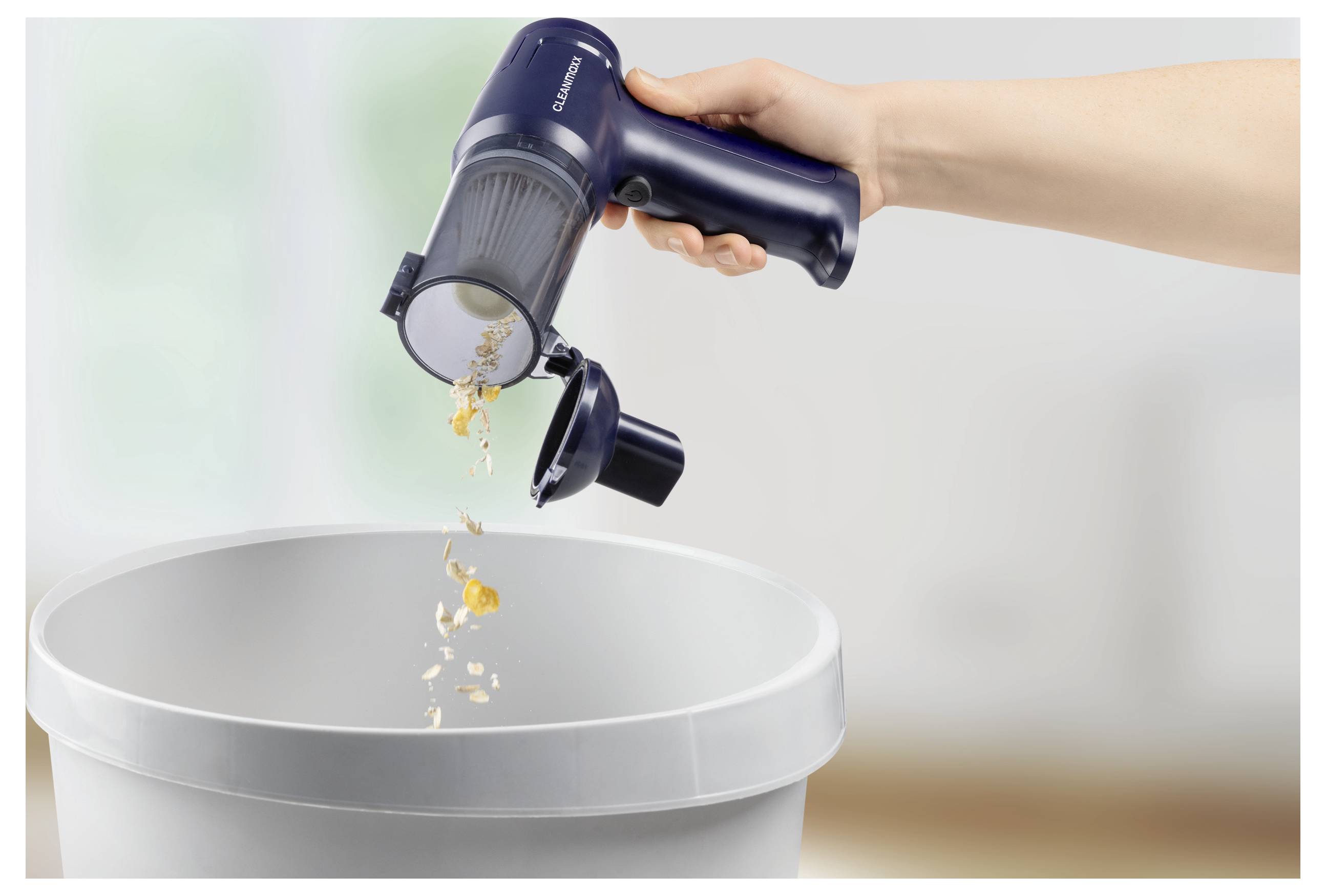 Eine Hand leert den Staubbehälter eines tragbaren Staubsaugers über einem Mülleimer aus, wobei Verschmutzungen herunterfallen. Die Szene spielt sich in einem Innenraum mit unscharfem Hintergrund ab.