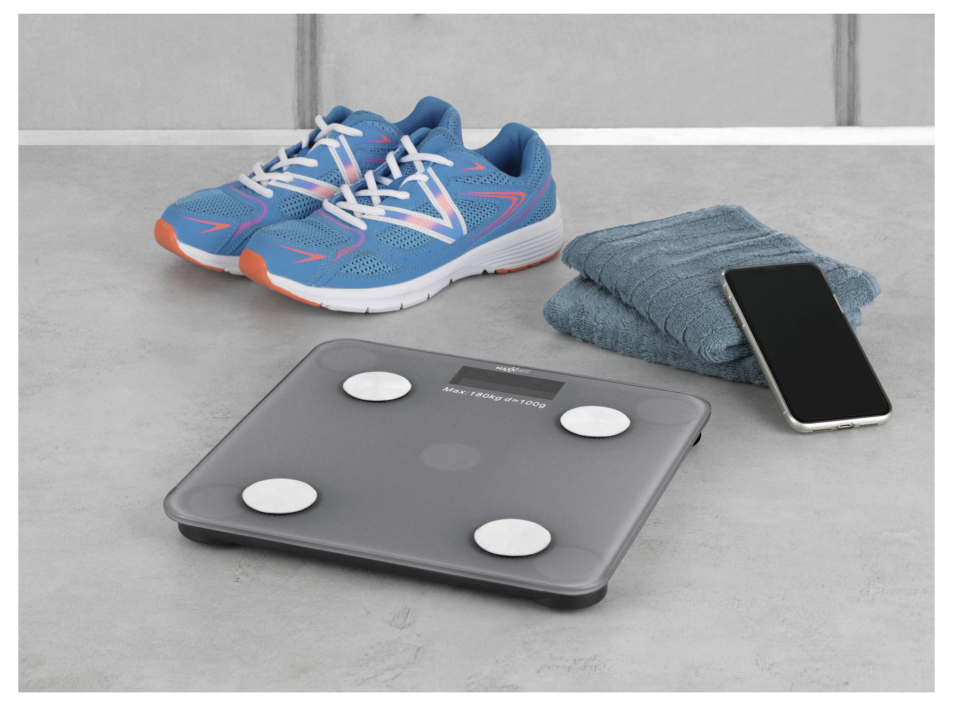 Eine digitale Badezimmerwaage neben einem Smartphone, blaue Sportschuhe und ein gefaltetes Handtuch auf einer grauen Oberfläche.