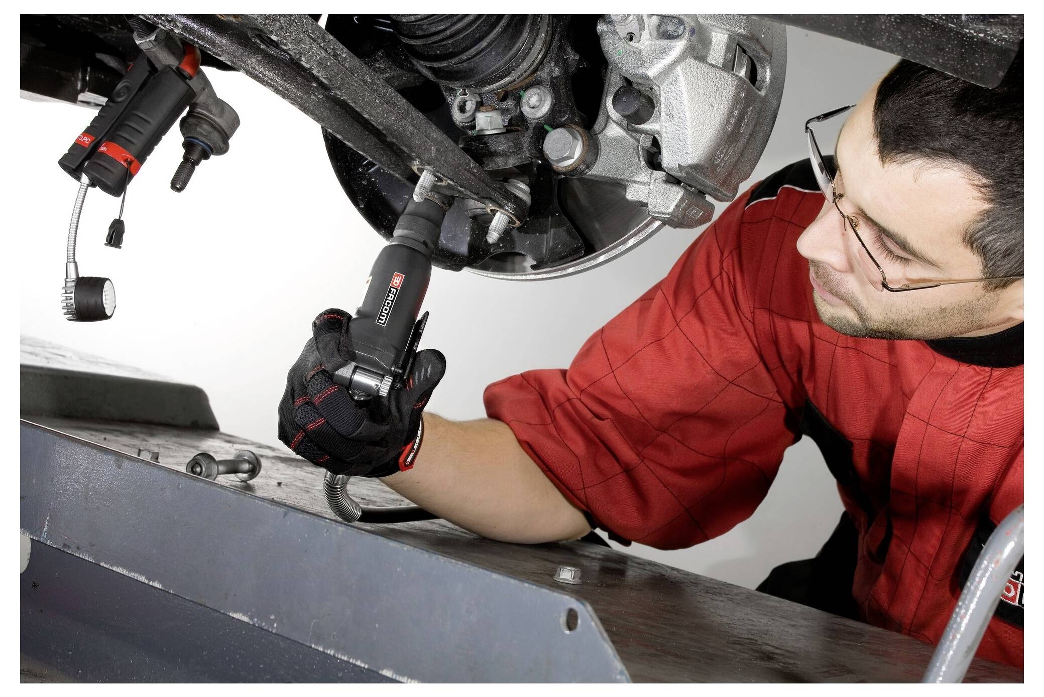 Ein Mechaniker in einem roten Hemd benutzt ein Elektrowerkzeug, um an der Unterseite eines Fahrzeugs zu arbeiten und konzentriert sich dabei auf eine Radnabe.
