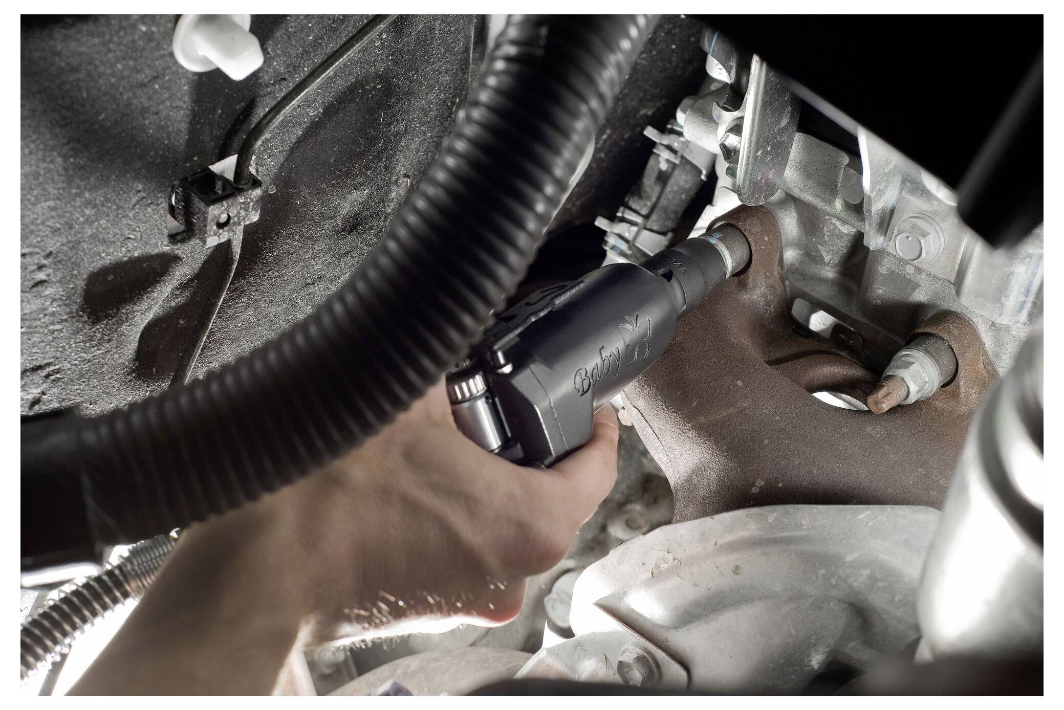 Eine Person verwendet einen Druckluftschrauber, um an der Unterseite eines Autos zu arbeiten, wobei sie sich auf einen rostigen Bolzen konzentriert, mit verschiedenen sichtbaren Fahrzeugkomponenten.
