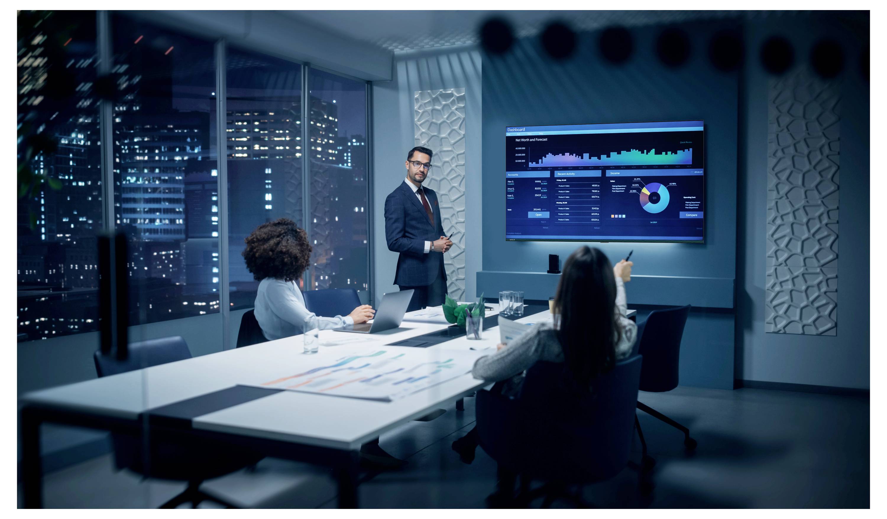 Geschäftstreffen in einem modernen Konferenzraum, bei dem ein Moderator Dashboards und Diagramme auf einem Wandbildschirm drei Kollegen zeigt, die um einen Tisch herum sitzen.