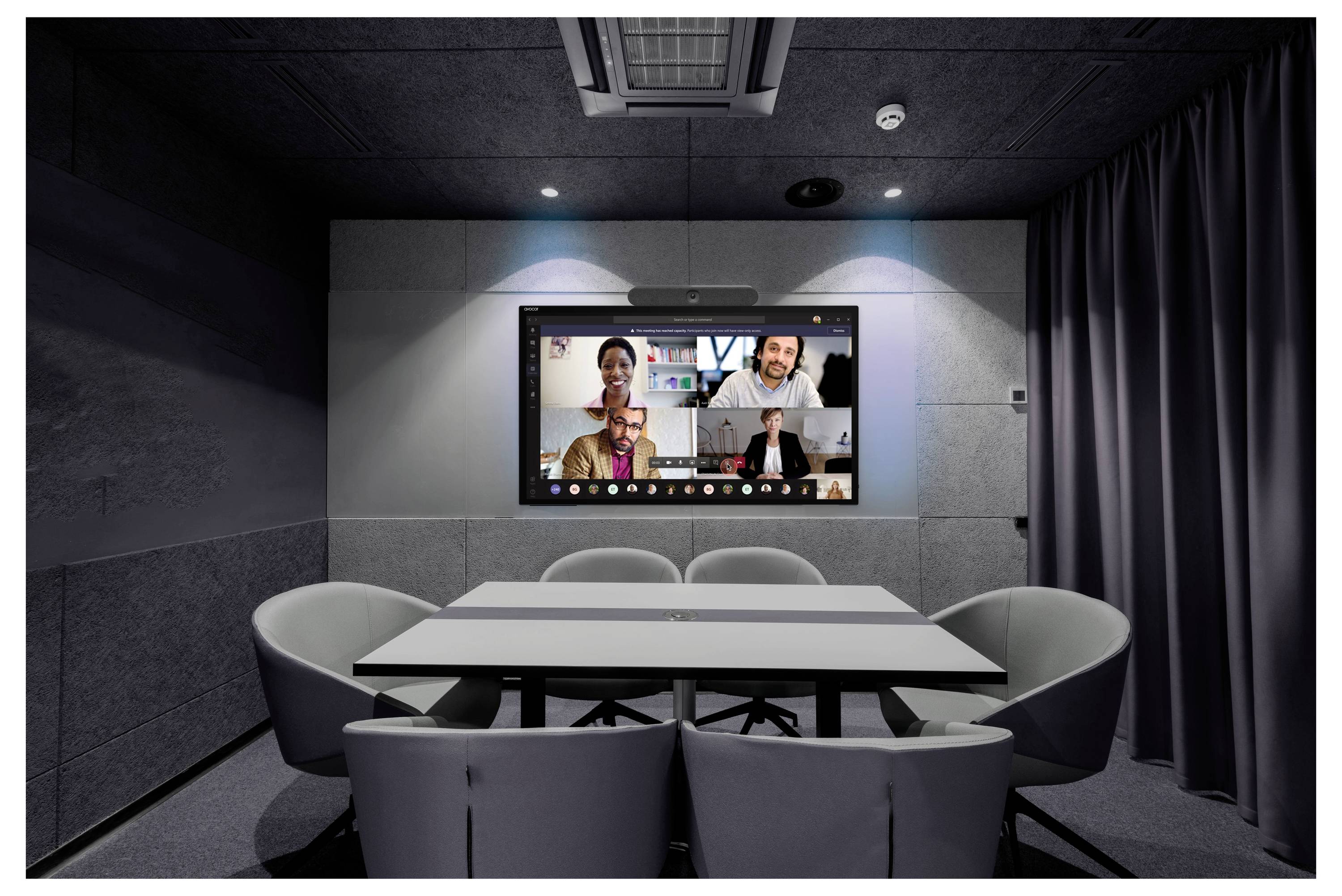 Kleiner moderner Konferenzraum mit einem an der Wand montierten Videobildschirm, auf dem vier Personen bei einer Videokonferenz um einen Tisch sitzen.