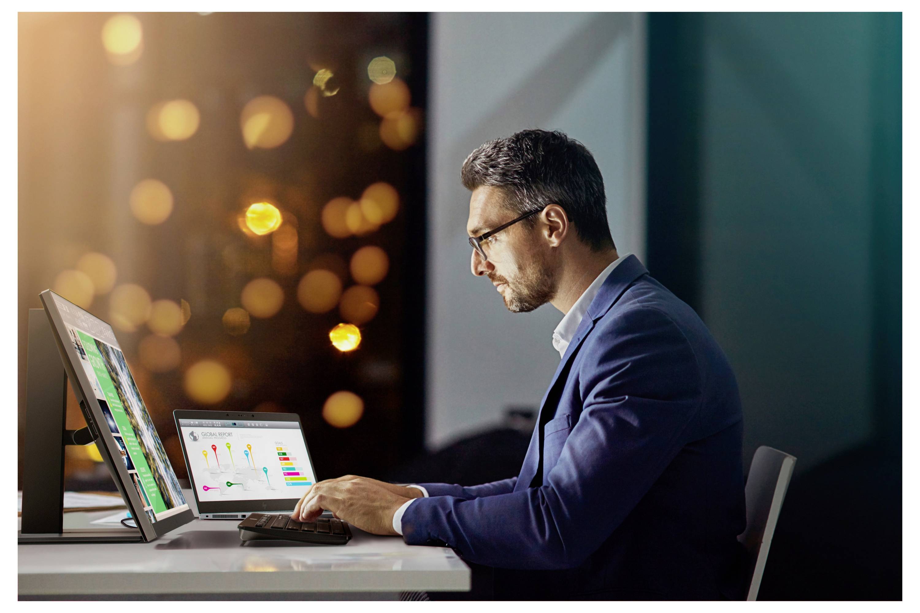 Ein Mann im Anzug arbeitet an einem Computer, auf dem Grafiken und Daten angezeigt werden. Der Hintergrund ist verschwommen mit sichtbaren Stadtlichtern.