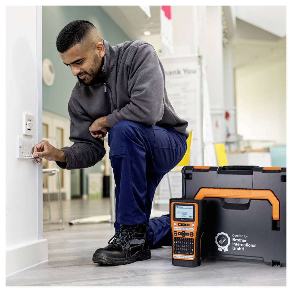 Techniker, der neben einer Wandsteckdose kniet und ein Prüfgerät verwendet; Werkzeugkoffer und Gerät neben ihm in einem Flur oder Büroraum.