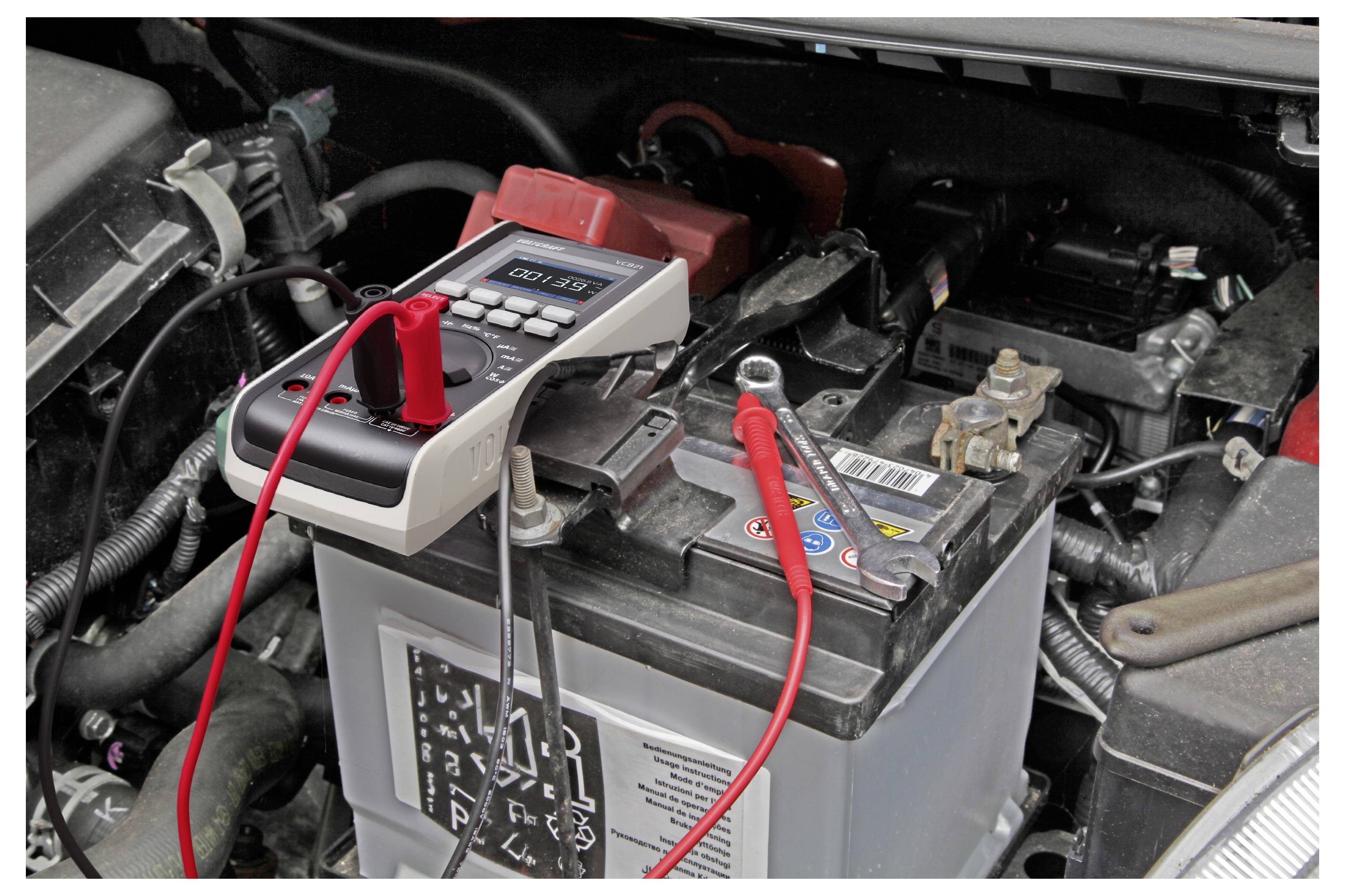 Ein digitales Multimeter mit roten und schwarzen Messspitzen prüft eine Autobatterie im Motorraum.
