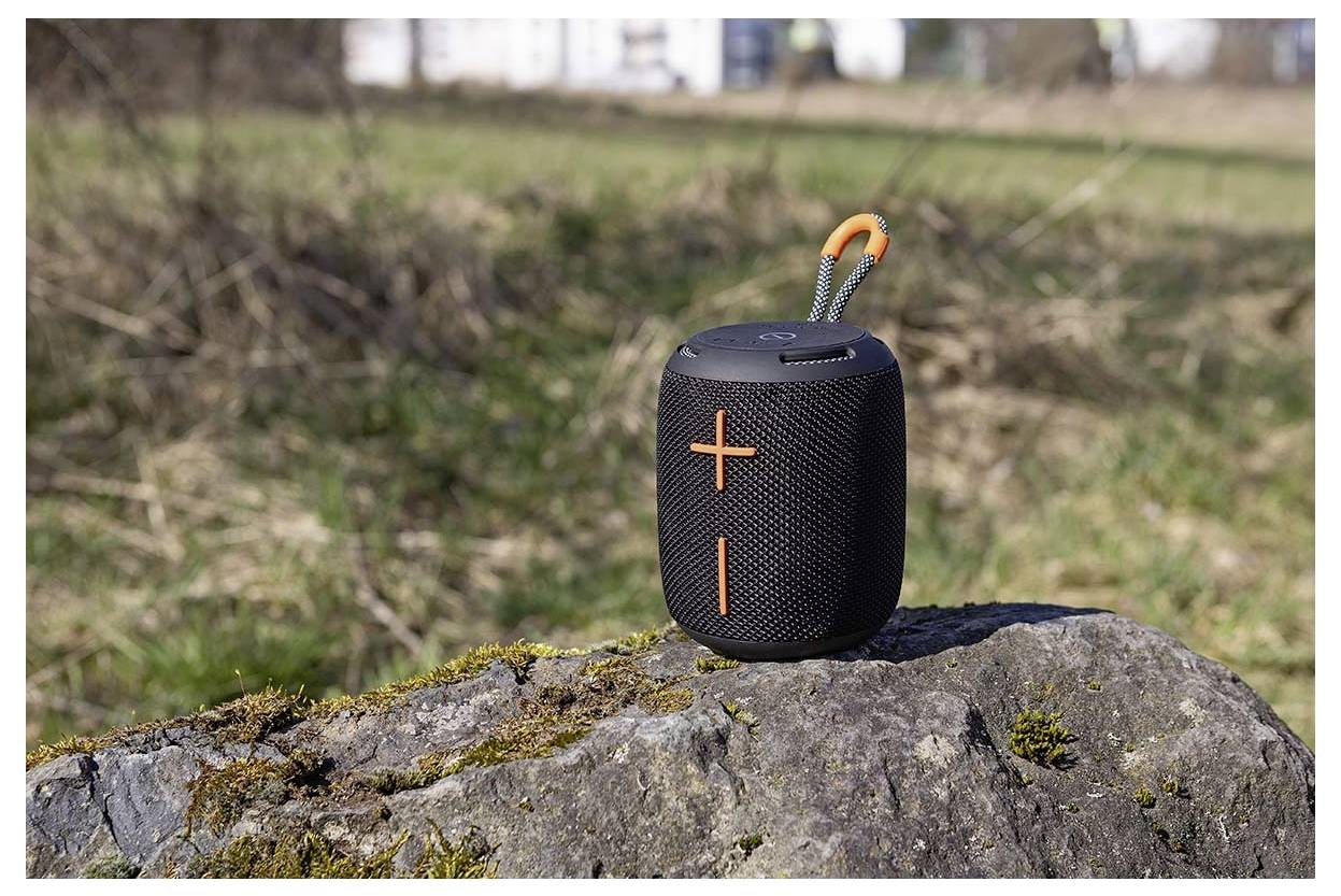 Ein tragbarer schwarzer und orangefarbener Bluetooth-Lautsprecher sitzt auf einem moosigen Stein in einer Außenumgebung mit unscharfem Gras im Hintergrund.