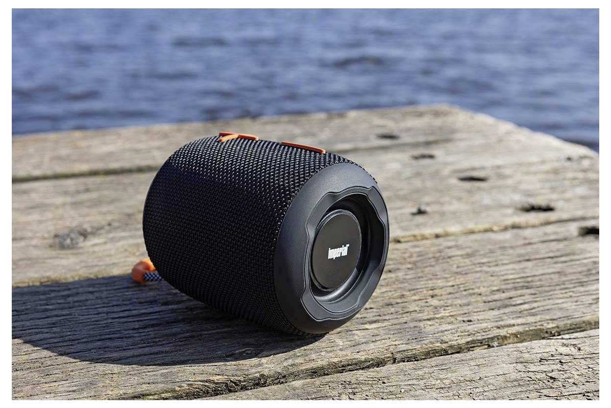 Ein tragbarer schwarzer Lautsprecher ruht auf einem hölzernen Steg an einem Gewässer und deutet auf die Nutzung im Freien und drahtlose Funktionalität hin.