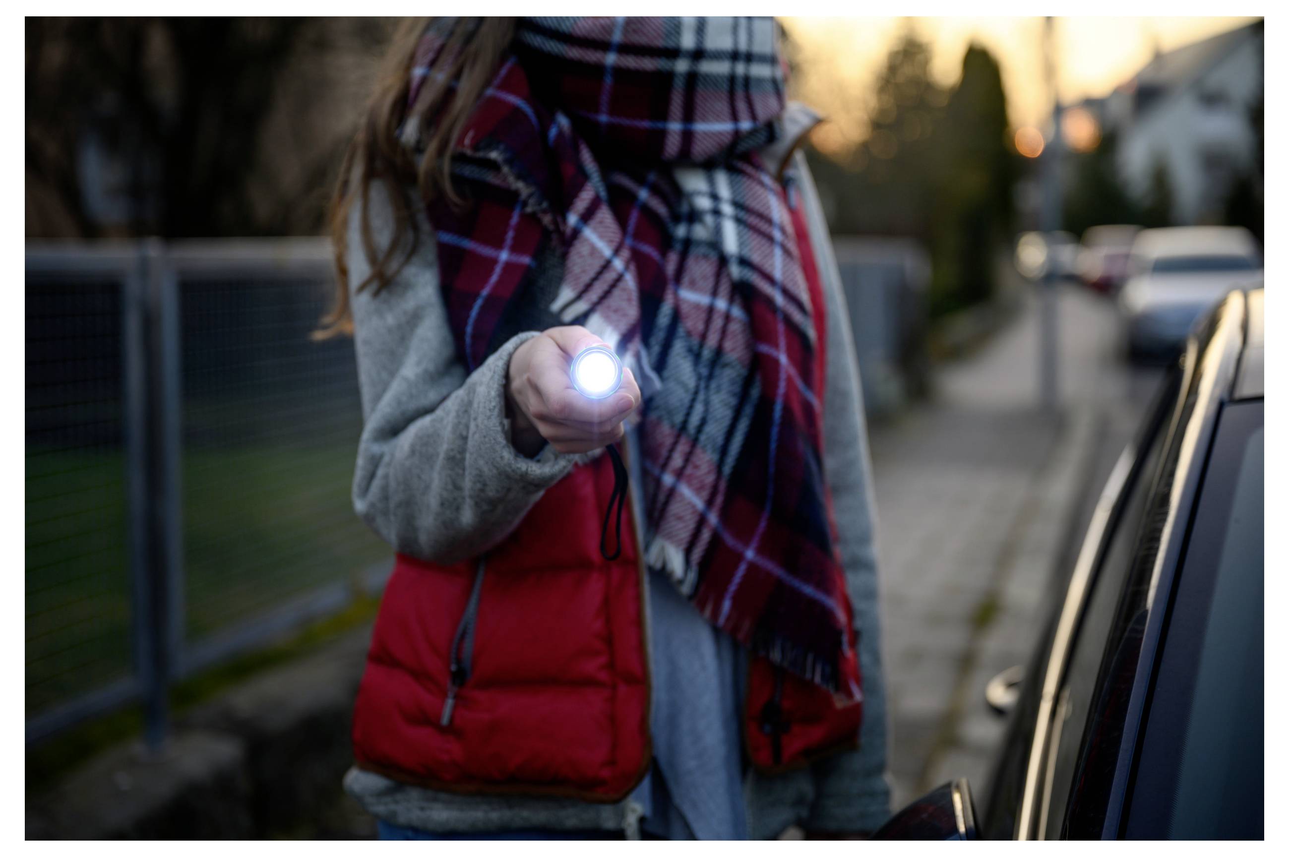 Person auf einem Gehweg, der eine Taschenlampe gegen ein Auto in der Dämmerung leuchtet, was darauf hindeutet, dass man in der Nähe eines geparkten Fahrzeugs überprüft oder signalisiert.