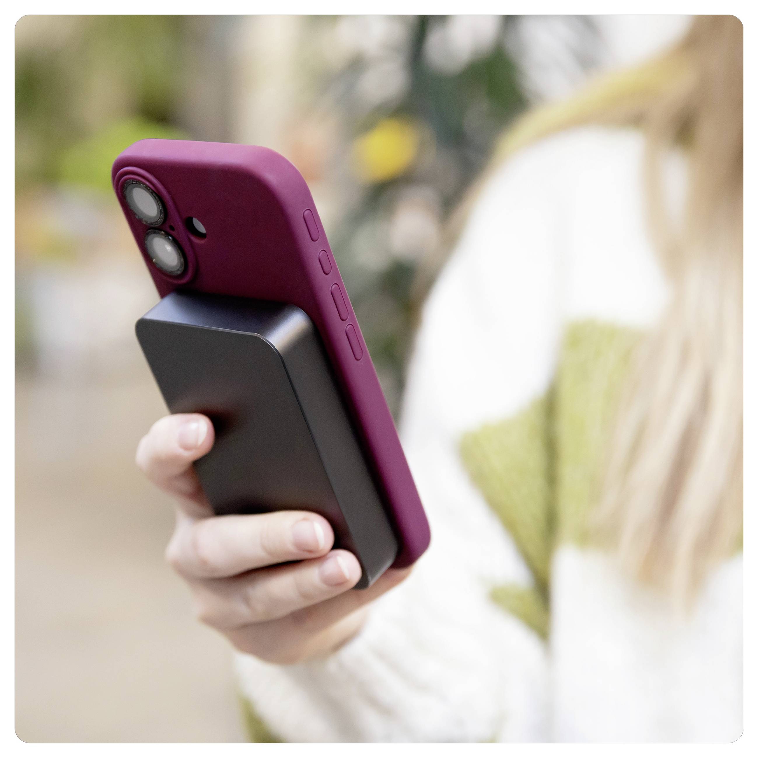 Hand, die ein Smartphone mit magentafarbenem Gehäuse und angebrachtem schwarzem Akkupack im Freien hält.