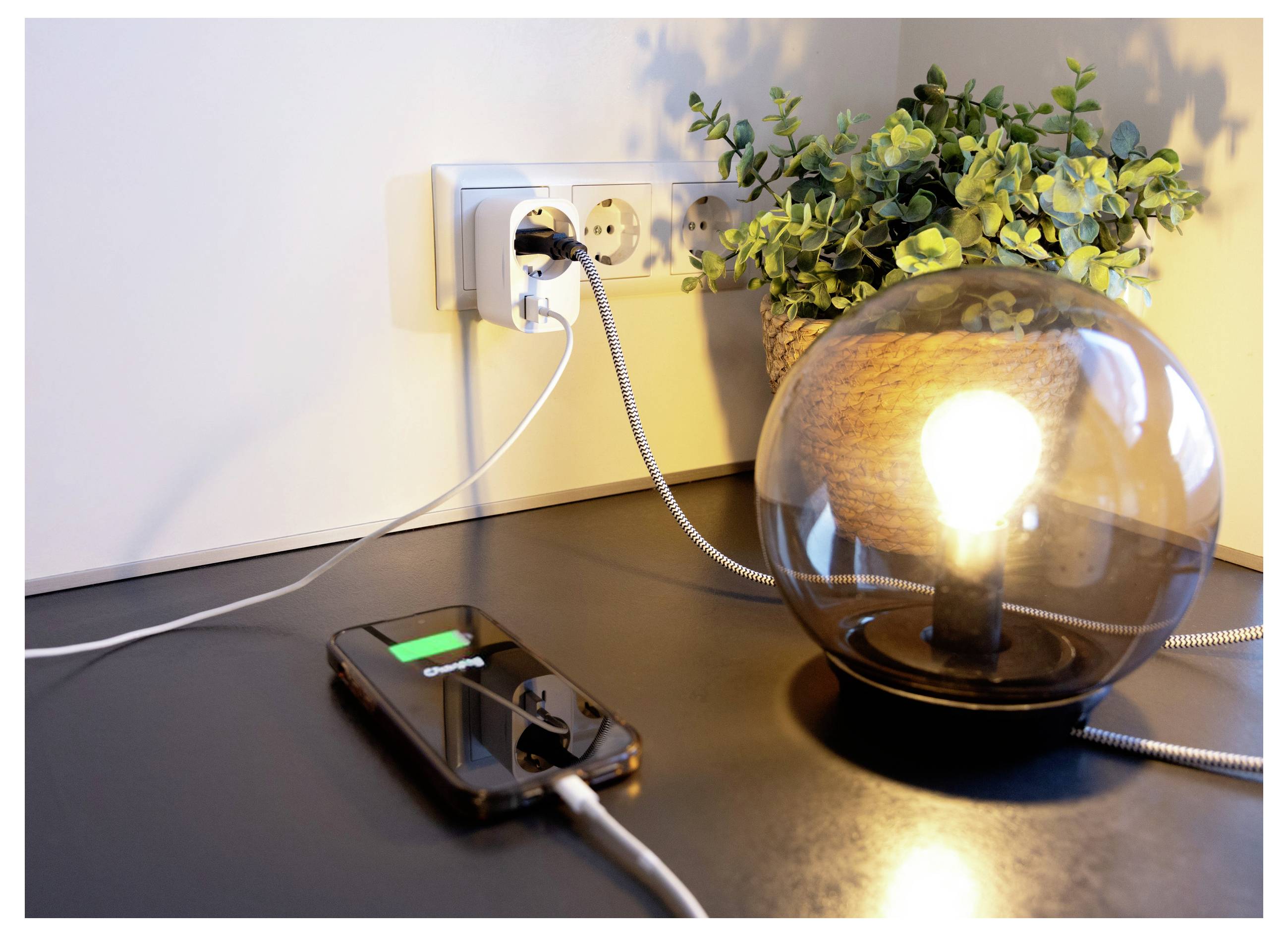 Smartphone wird auf einem Schreibtisch neben einer leuchtenden Tischlampe aufgeladen, die in eine Wandsteckdose eingestöpselt ist, mit einer Topfpflanze im Hintergrund.