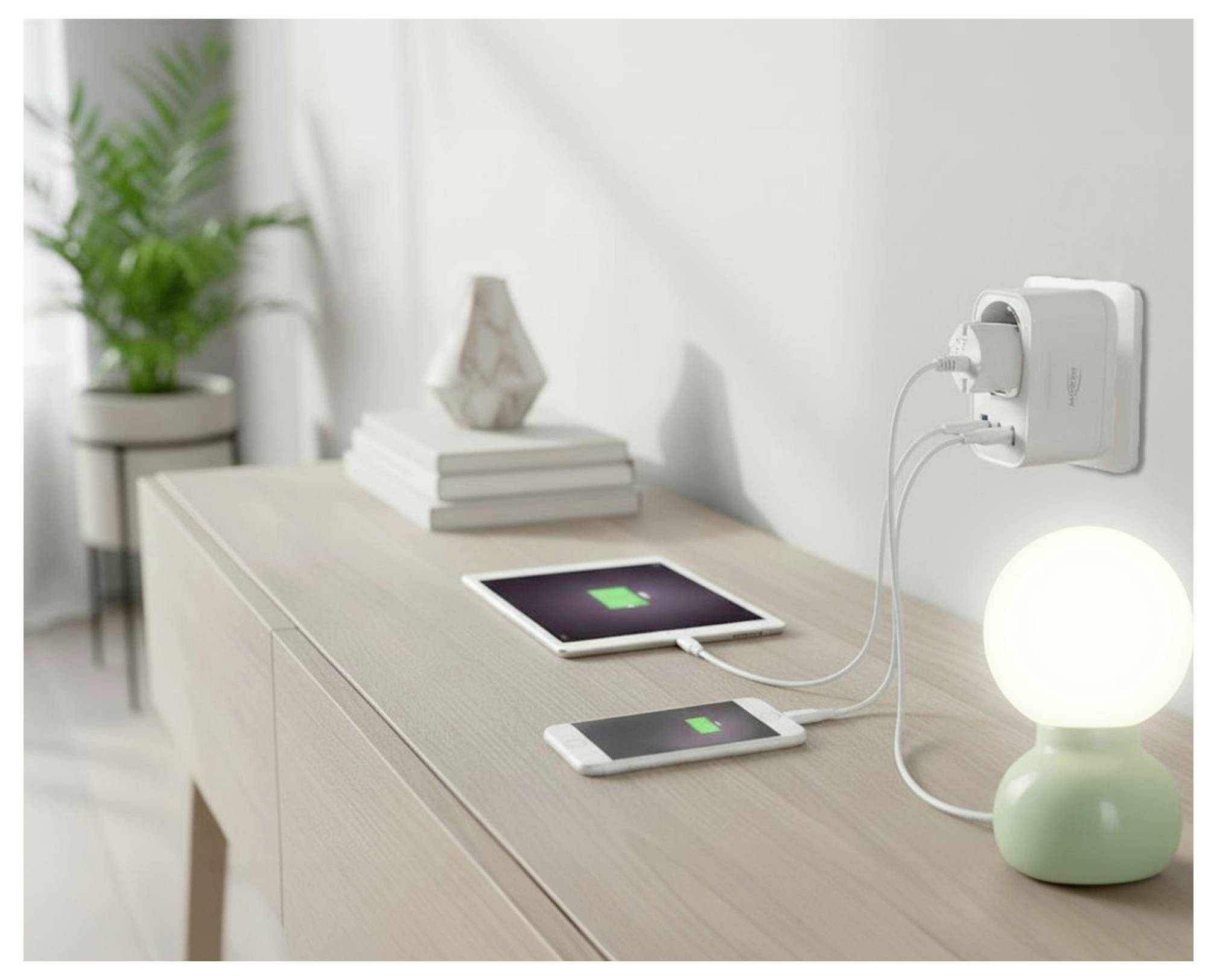 Zwei Geräte, die von einer Steckdose an der Wand neben einer leuchtenden Tischlampe und gestapelten Büchern auf einem Schreibtisch aufgeladen werden.
