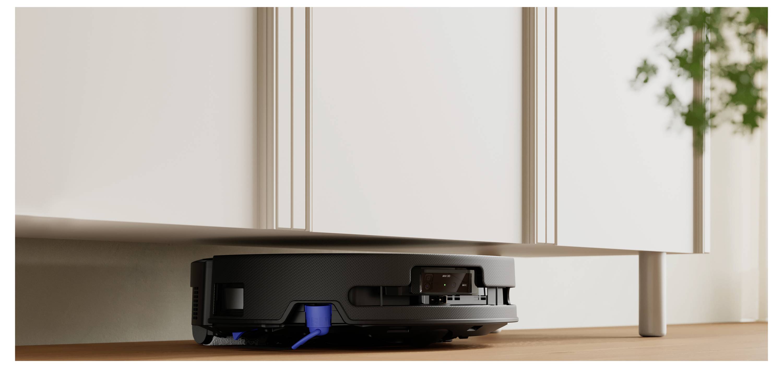 Ein schwarzer robotischer Staubsauger navigiert unter einem weißen Schrank auf einem Holzboden, während eine Pflanze im Hintergrund sichtbar ist.