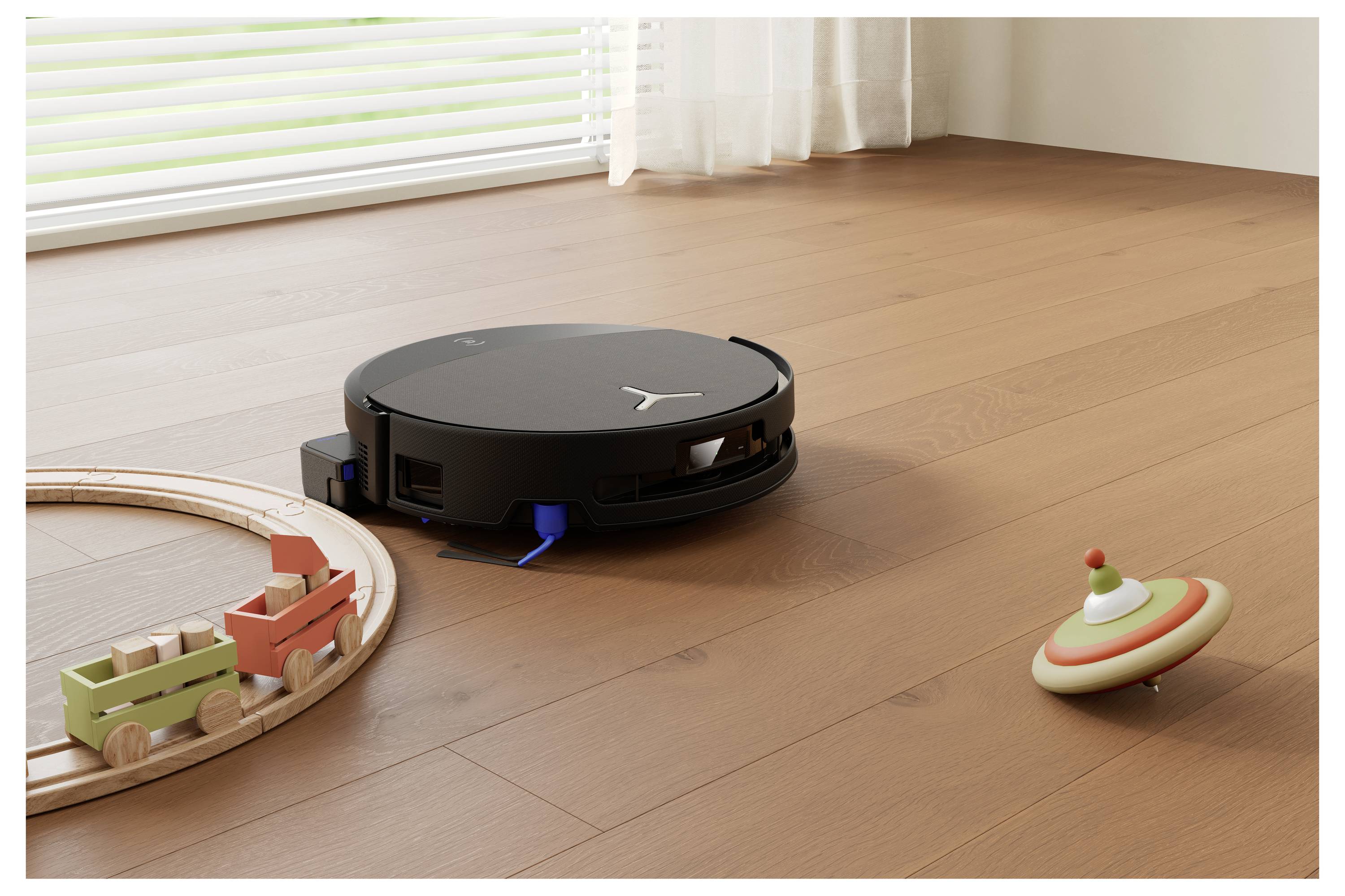 Ein schwarzer kreisförmiger Roboterstaubsauger reinigt einen Holzboden in der Nähe einer Spielzeugeisenbahn auf Gleisen und eines Kreisels in einem sonnigen Zimmer.