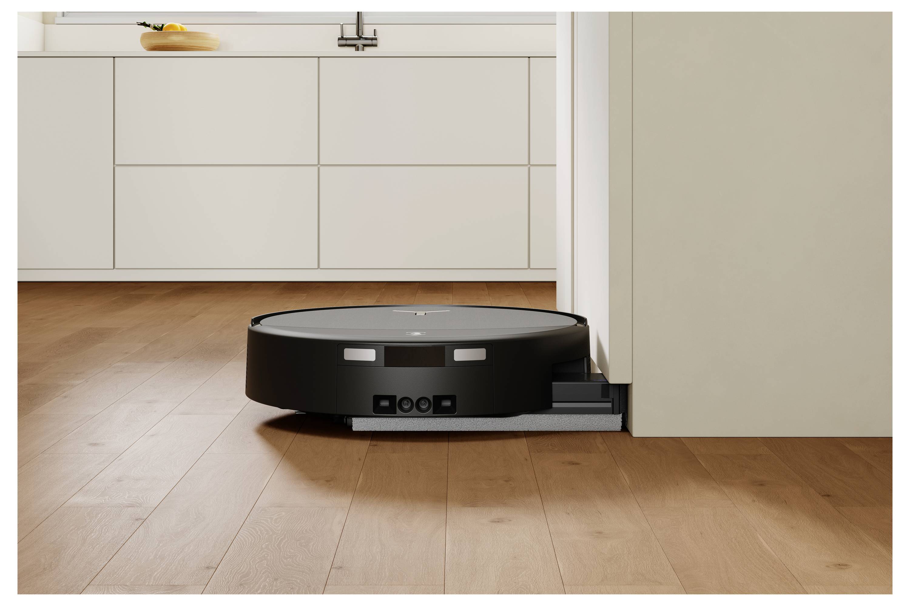 Ein schwarzer Roboterstaubsauger dockt sich an einer weißen Sockelleiste in einem Zimmer mit Holzböden an.