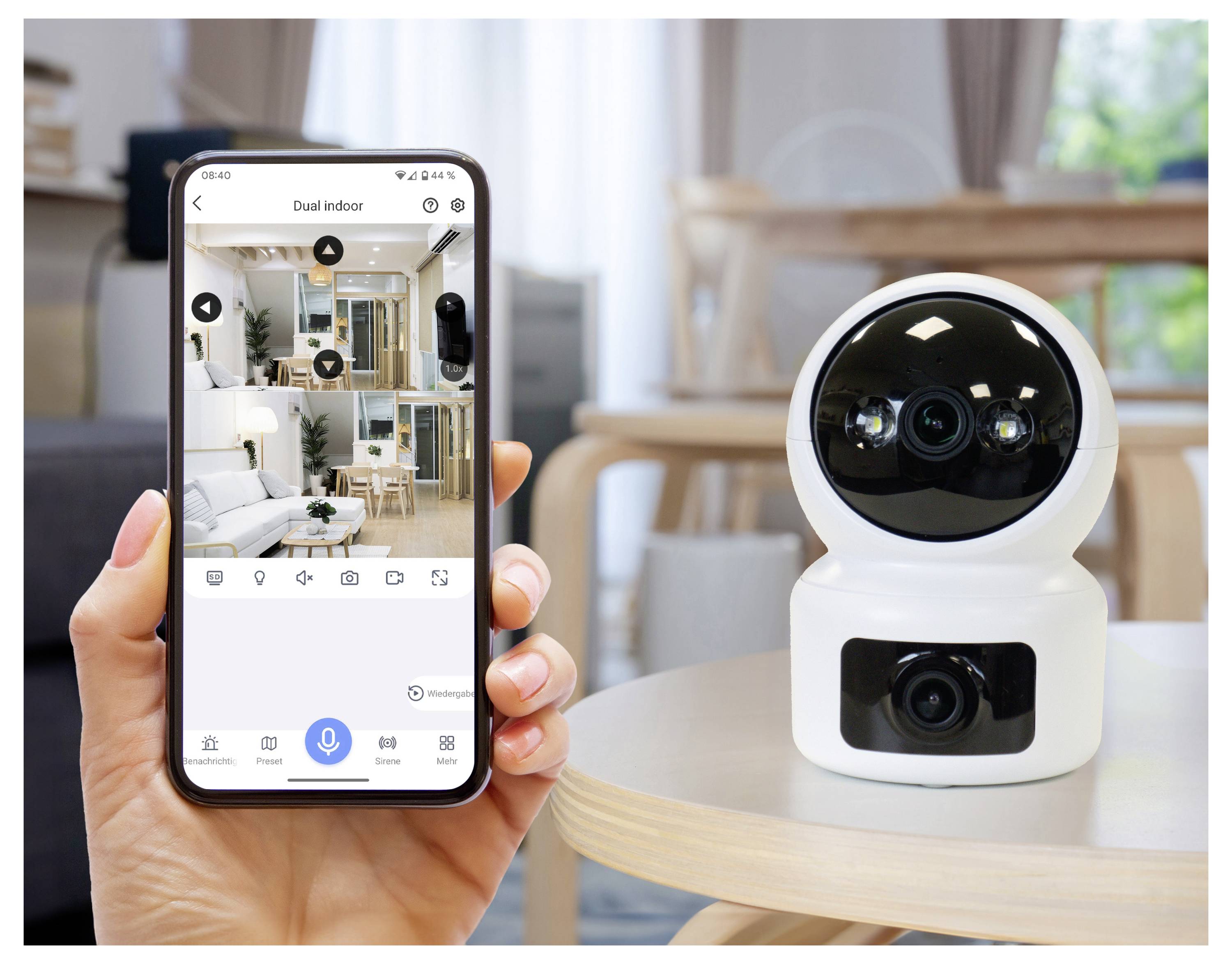 Eine Person hält ein Smartphone, auf dem eine Hausautomations-App angezeigt wird, die die Ansicht einer Dual-Innenkamera zeigt. Die Kamera steht auf einem nahegelegenen Tisch.