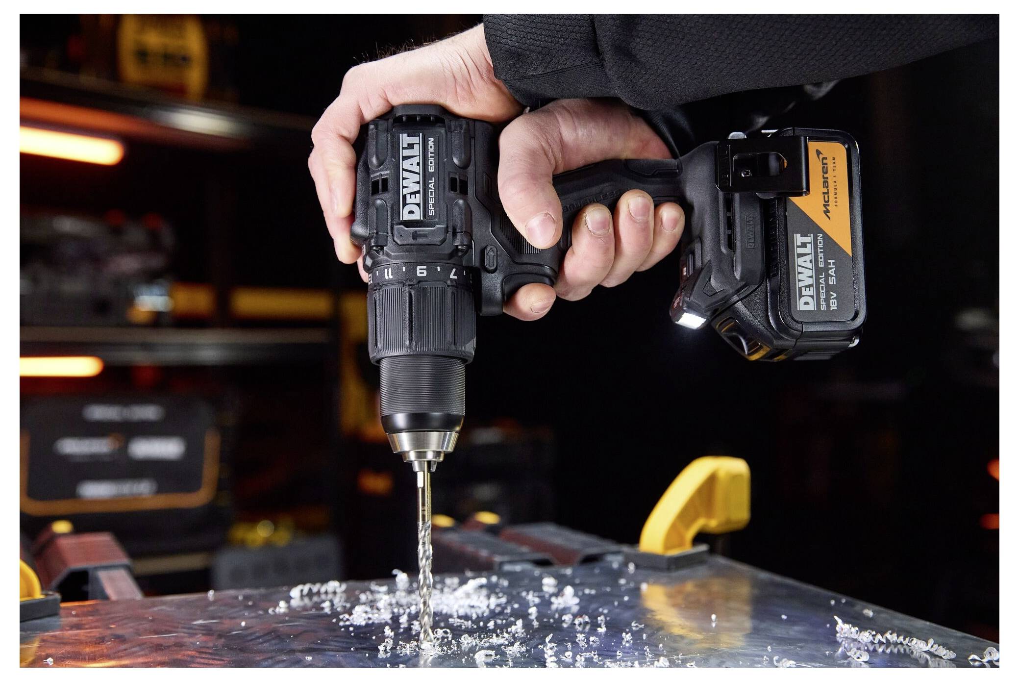 Hand hält einen DEWALT Akku-Bohrer mit Bit, der in Metall bohrt und Späne erzeugt; Akkupack des Werkzeugs sichtbar in einer Werkstattumgebung.