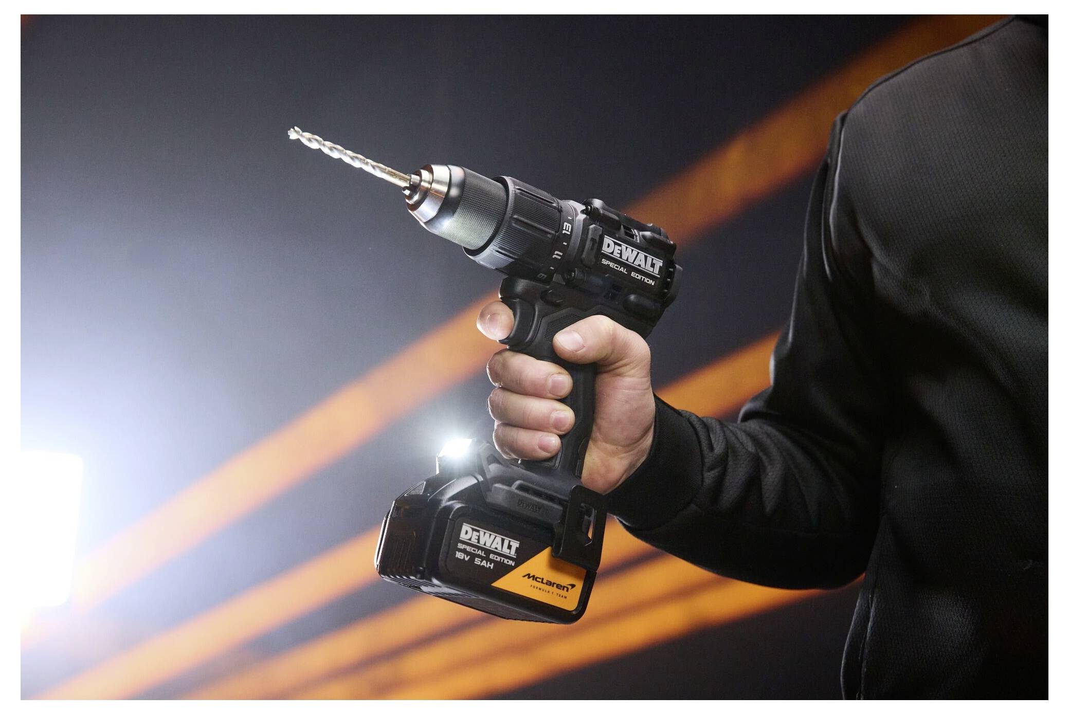 Person, die eine DEWALT Akku-Bohrmaschine mit einem Bohraufsatz hält, vor einem dunklen Hintergrund mit orangefarbenen Lichtstreifen abgebildet.