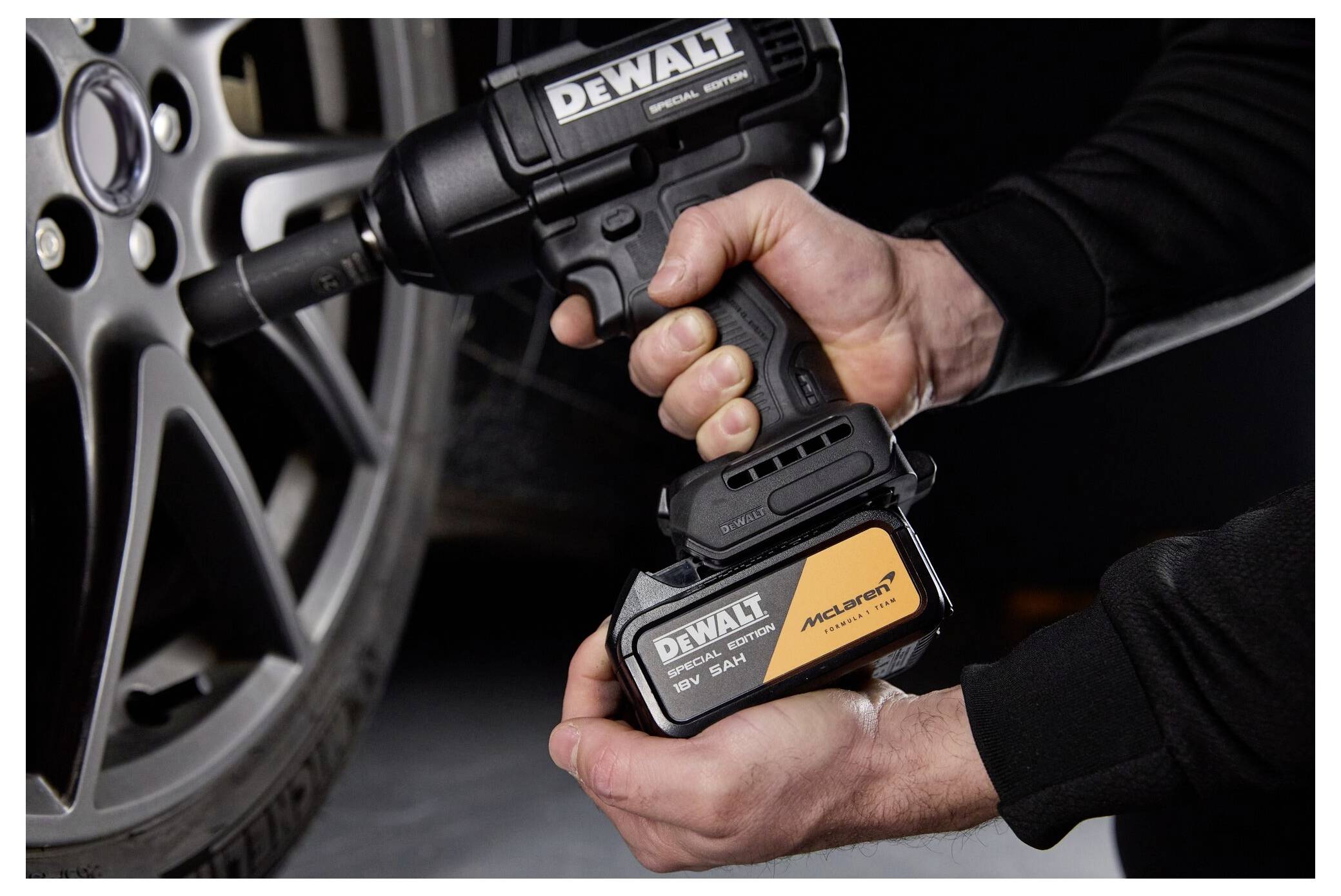 Hände halten einen DEWALT kabellosen Schlagschrauber und ein Akkupack neben einem Autoreifen und zeigen die Installation des Werkzeugs und des Akkus.