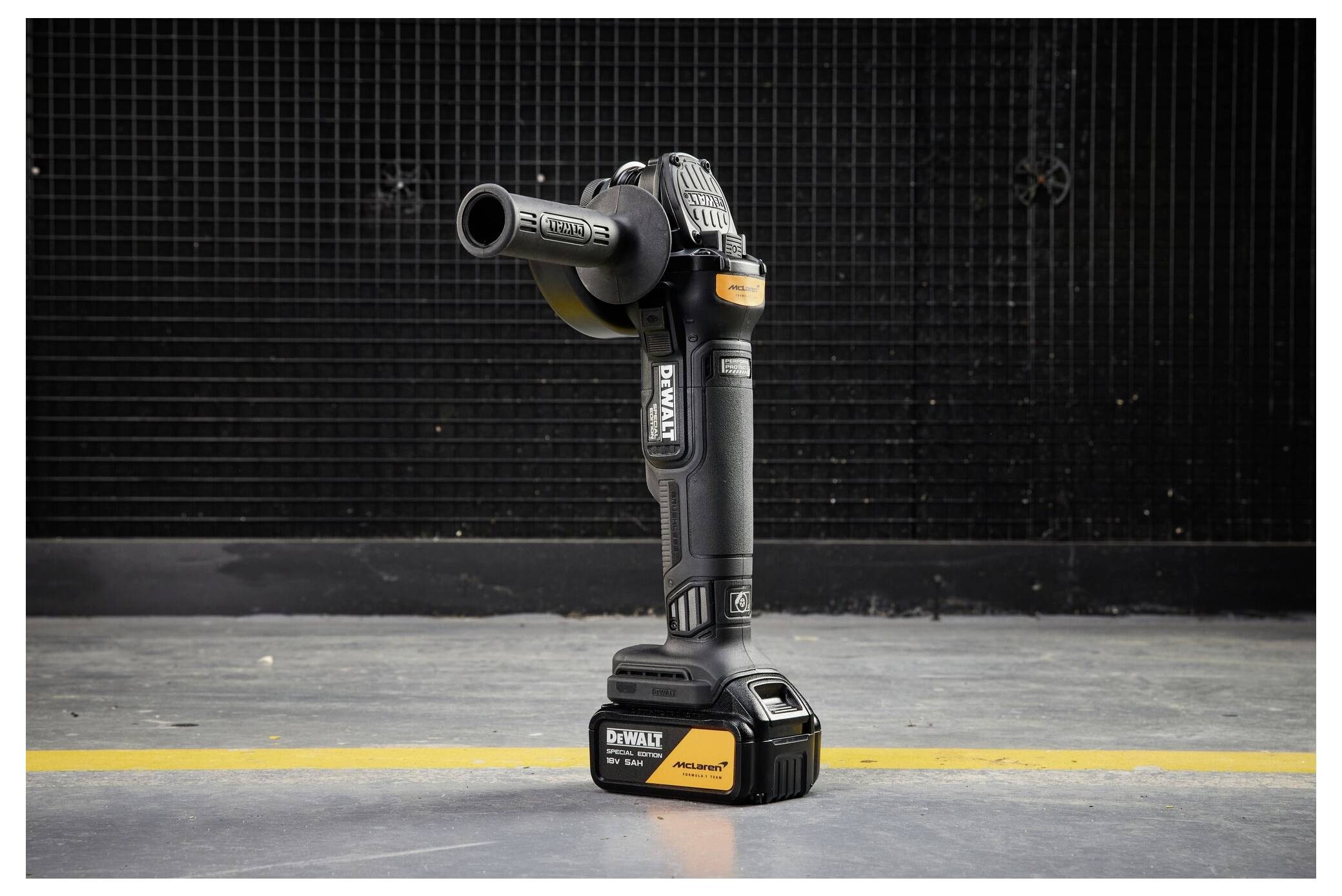 Ein DeWalt-Akkuschleifers steht aufrecht auf einem Betonboden vor einer dunklen Wand, wobei der Seitengriff angebracht ist.