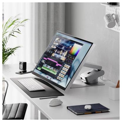 Moderner Schreibtischaufbau mit einem großen geneigten Monitor, der Videobearbeitungssoftware anzeigt, Tastatur, Maus, Notizbuch und einem kleinen Desktop-Scanner in einem hellen Arbeitsbereich.