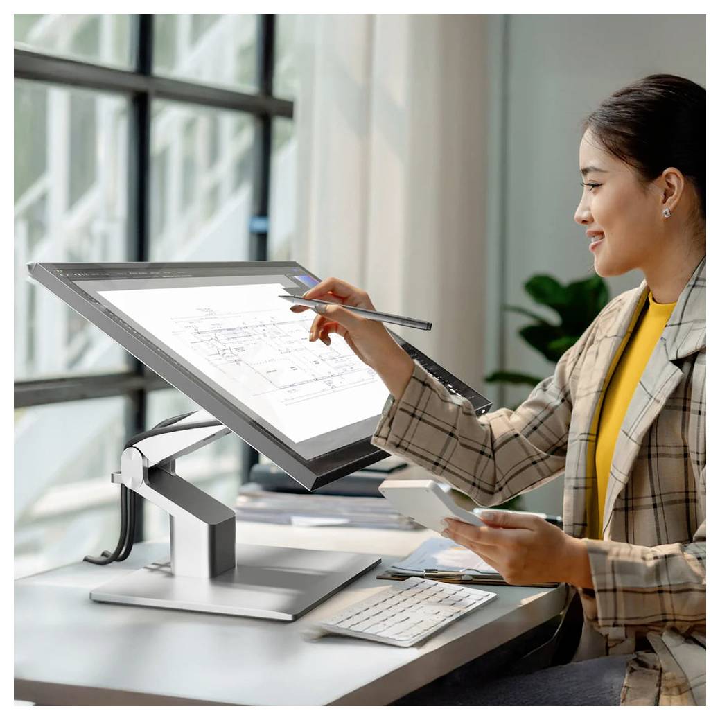 Frau überprüft eine technische Zeichnung auf einem Pen-Display an einem Schreibtisch, hält einen Stylus und ein Telefon in einem modernen Büro.
