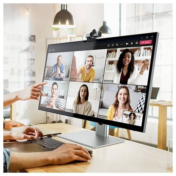 Eine Person nimmt an einer Videokonferenz auf einem Desktopmonitor teil, der sechs lächelnde Teilnehmer in einem Rasterformat zeigt, mit einer angebrachten Webcam und einer Tastatur auf dem Schreibtisch.
