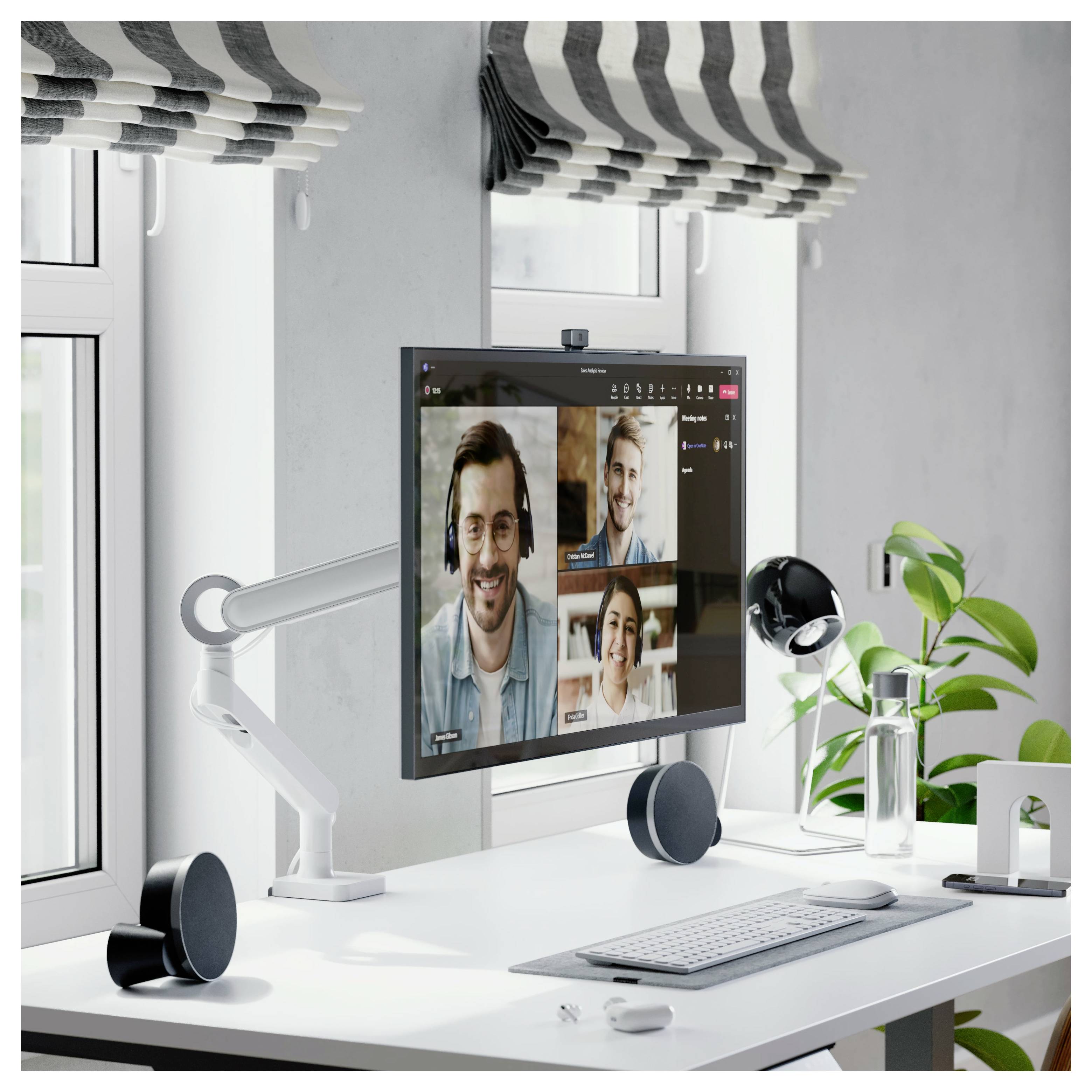 Modernes Homeoffice mit einem Monitor, auf dem ein Videoanruf mit drei Personen angezeigt wird, sowie Tastatur, Lautsprecher, Lampe und eine Topfpflanze auf einem weißen Schreibtisch.