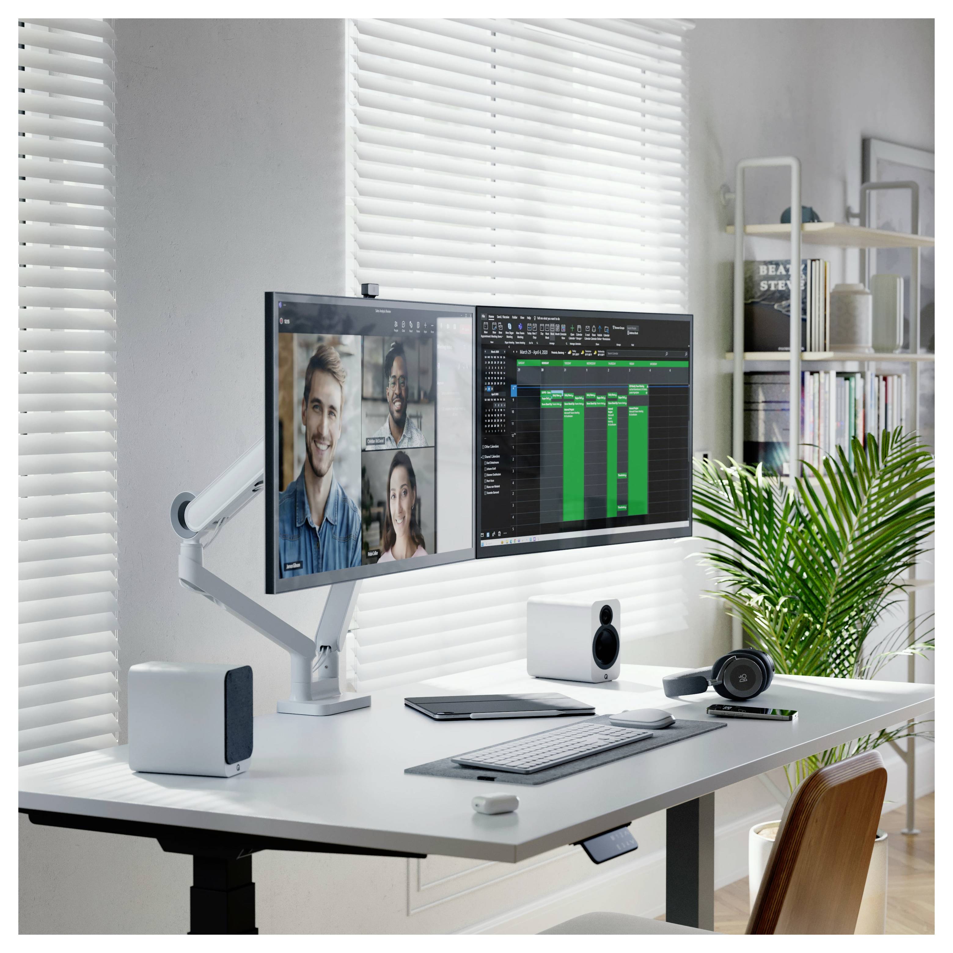 Moderner Schreibtisch im Homeoffice mit zwei Monitoren, auf denen ein Videoanruf und ein Projektzeitplan angezeigt werden, sowie eine Tastatur, Lautsprecher und eine Pflanze neben einem hellen Fenster.