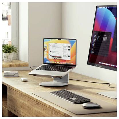 Modernes Schreibtisch-Setup mit einem Laptop auf einem Ständer, einem externen Monitor, einer Tastatur und einer Maus auf einem Holzarbeitsplatz neben einem Fenster.