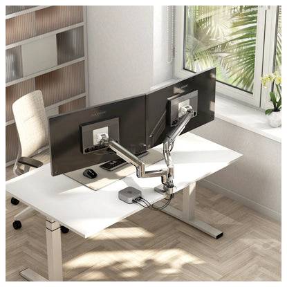 Moderner Büroschreibtisch mit zwei Monitoren auf beweglichen Armen, Tastatur, Maus und Stuhl neben einem Fenster in einem hellen Arbeitsbereich.