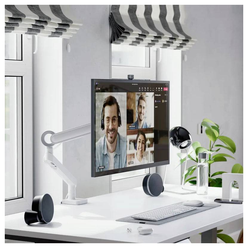 Homeoffice-Schreibtisch mit Monitor, auf dem ein Videoanruf mit drei Personen zu sehen ist, Tastatur, Kopfhörer, Schreibtischlampe und Topfpflanze am Fenster.