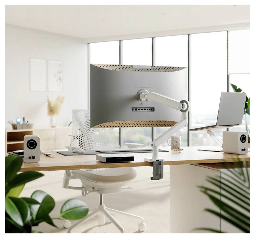 Modernes Homeoffice mit großem gebogenem Monitor auf einem verstellbaren Arm, Laptop, Schreibtischlautsprechern, Tastatur und Stuhl neben hellen Fenstern.