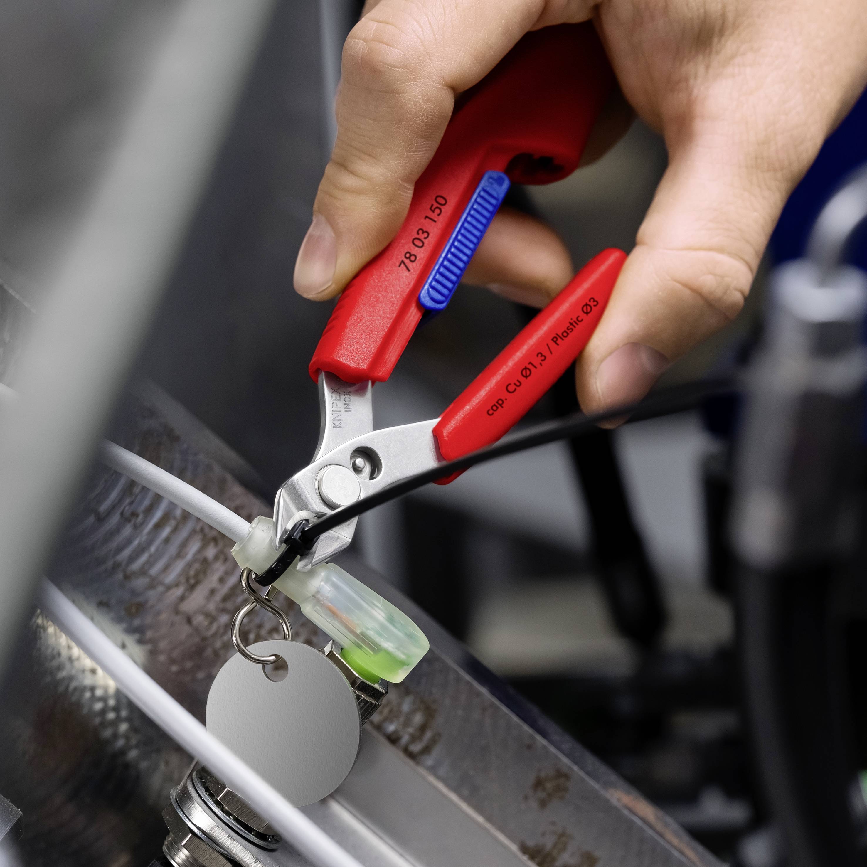 Hand, die rote Seitenschneider verwendet, um einen Kabelbinder zu durchtrennen, der einen Schlüsselring an einem Metallrahmen befestigt.