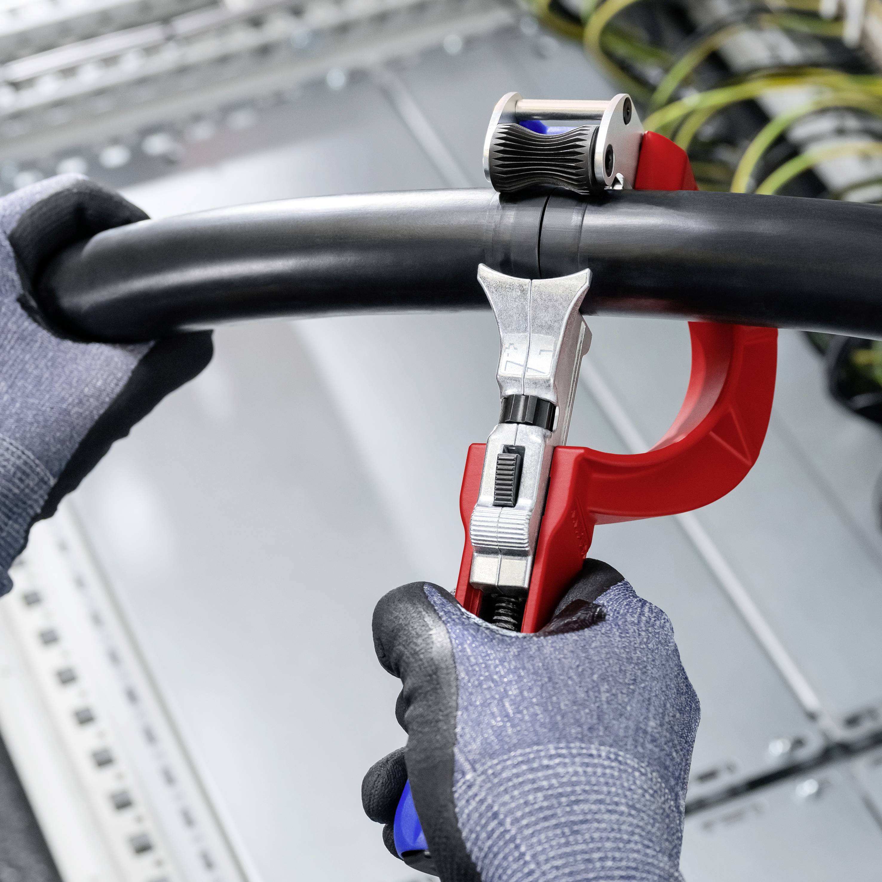 Handschuhe tragenden Hände verwenden einen roten Kabelschneider, um ein dickes schwarzes Kabel in einer Industrieumgebung zu durchtrennen.