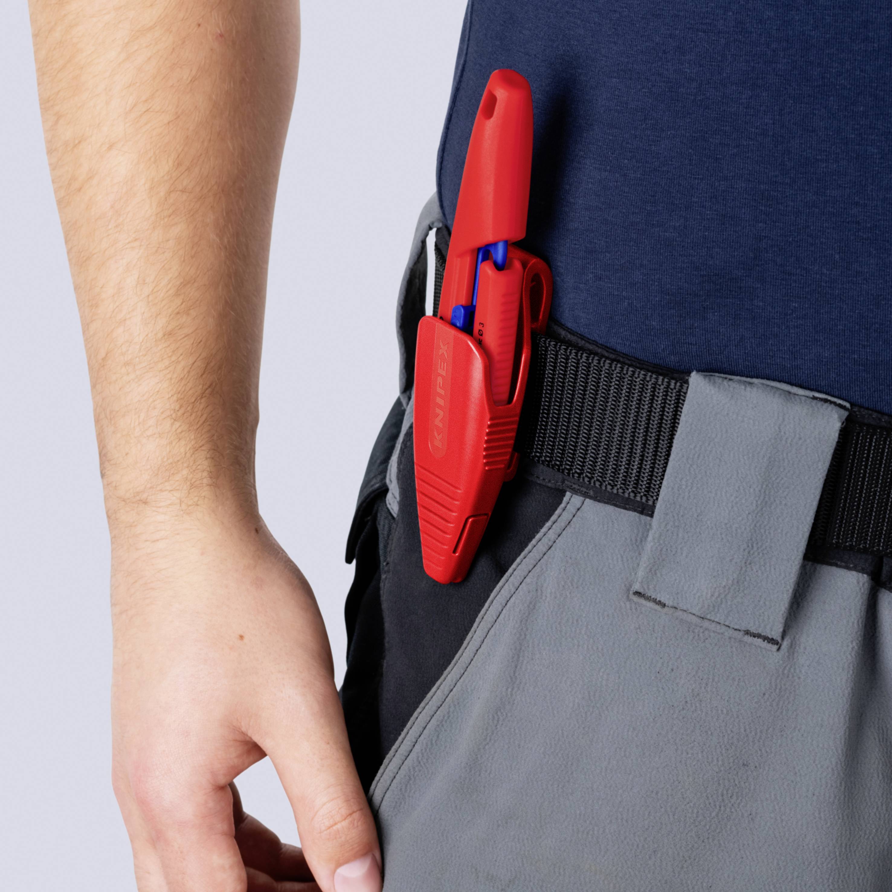Nahaufnahme einer Person, die ein rotes Universalmesser an einem grauen Arbeitsgürtel neben ihrer Hand befestigt trägt.