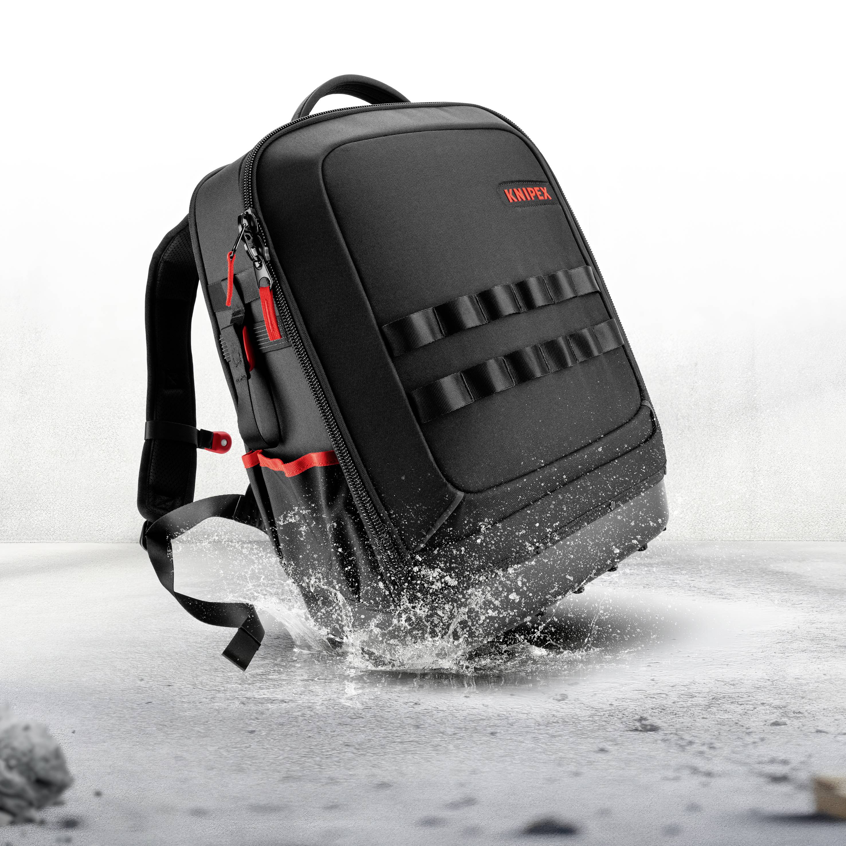 Schwarzer KNIPR-Rucksack mit roten Akzenten, der durch Wasser spritzt, aufrecht auf einer weißen Oberfläche stehend dargestellt.