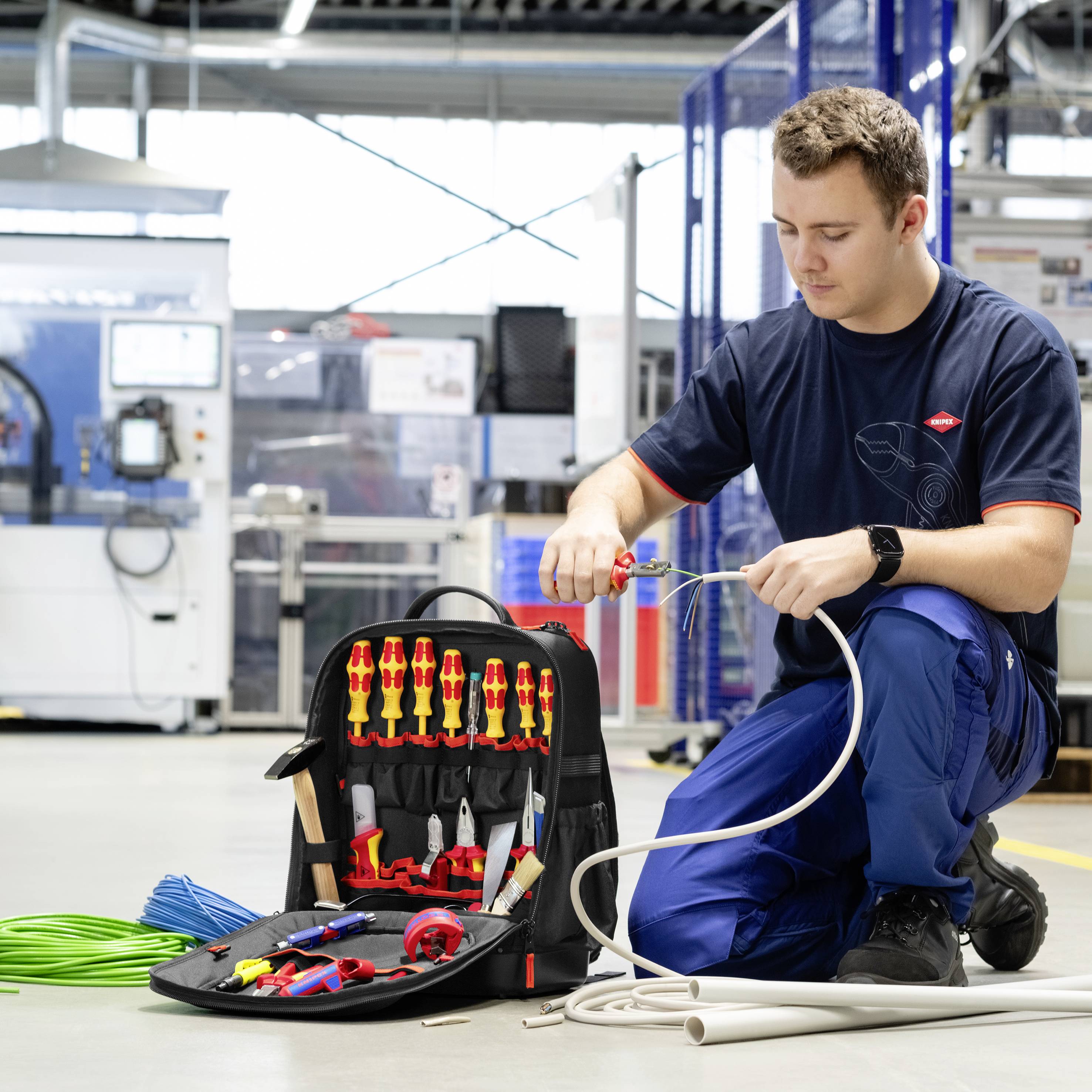 Techniker kniet in einer Werkstatt und testet sowie verdrahtet Kabel neben einer offenen Werkzeugtasche, die mit Zangen, Schneidern und isolierten Schraubendrehern gefüllt ist.